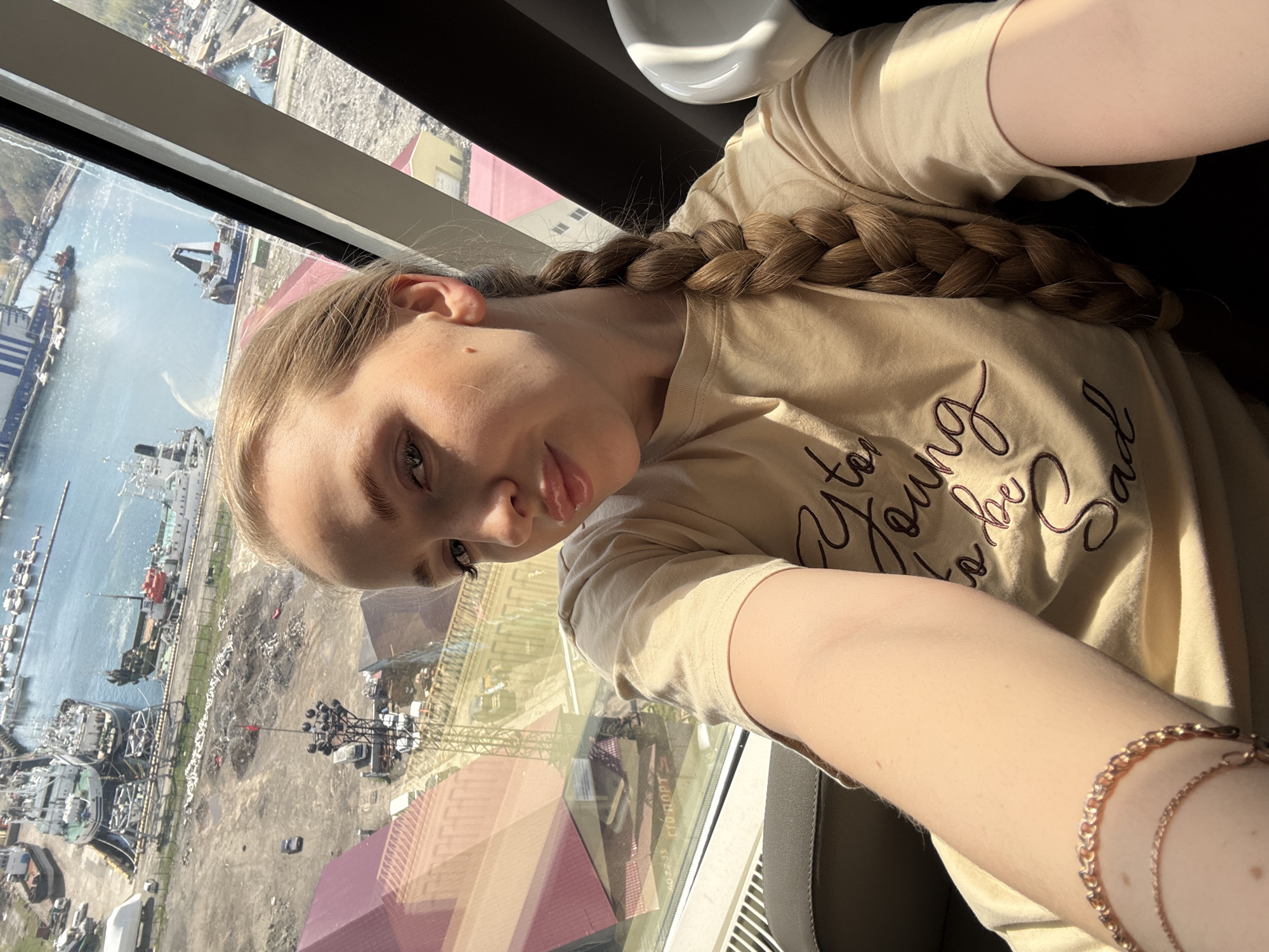 The image is a rotated selfie of a young woman, taken from an elevated perspective overlooking an industrial port. The woman has long, light-colored hair styled in a thick braid that rests over her shoulder. She is wearing a light beige t-shirt with the partially visible text "Too Young to be Sad" embroidered or printed on the front. A rose gold-toned bracelet adorns her right wrist. She gazes directly at the camera with a neutral expression.
Through a large window, a bustling industrial port or harbor is visible under bright, clear daylight, consistent with the provided location context of Petropavlovsk-Kamchatsky, Russia. The sunlit water reflects the clear sky. Numerous ships of various sizes, including cargo vessels and smaller boats, are docked at extensive piers or moving through the water. The surrounding land features industrial structures, cranes, and storage areas. Several buildings with reddish roofs can be seen in the mid-ground. Sunlight streams into the room, creating highlights on the woman's hair and arm. Reflections on the window glass reveal some text from external elements, including "СТО ПОРТ" (STO PORT) and the numbers "2022-33". The scene depicts an active, industrious coastal environment on a sunny day.