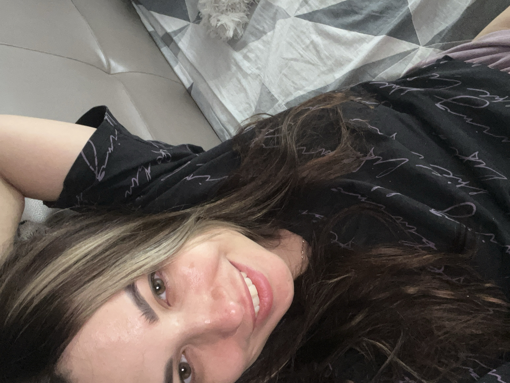 A young woman is lying on a light grey leather-like surface. She is wearing a black shirt with light purple script. Her hair is long, dark brown with blonde highlights. She is smiling and looking at the camera. Behind her is a bed with a grey and white geometric patterned comforter. A small, white, fluffy object is on the comforter. The location is an indoor space, likely a residential bedroom or living room. The city and state are Unknown, Unknown.