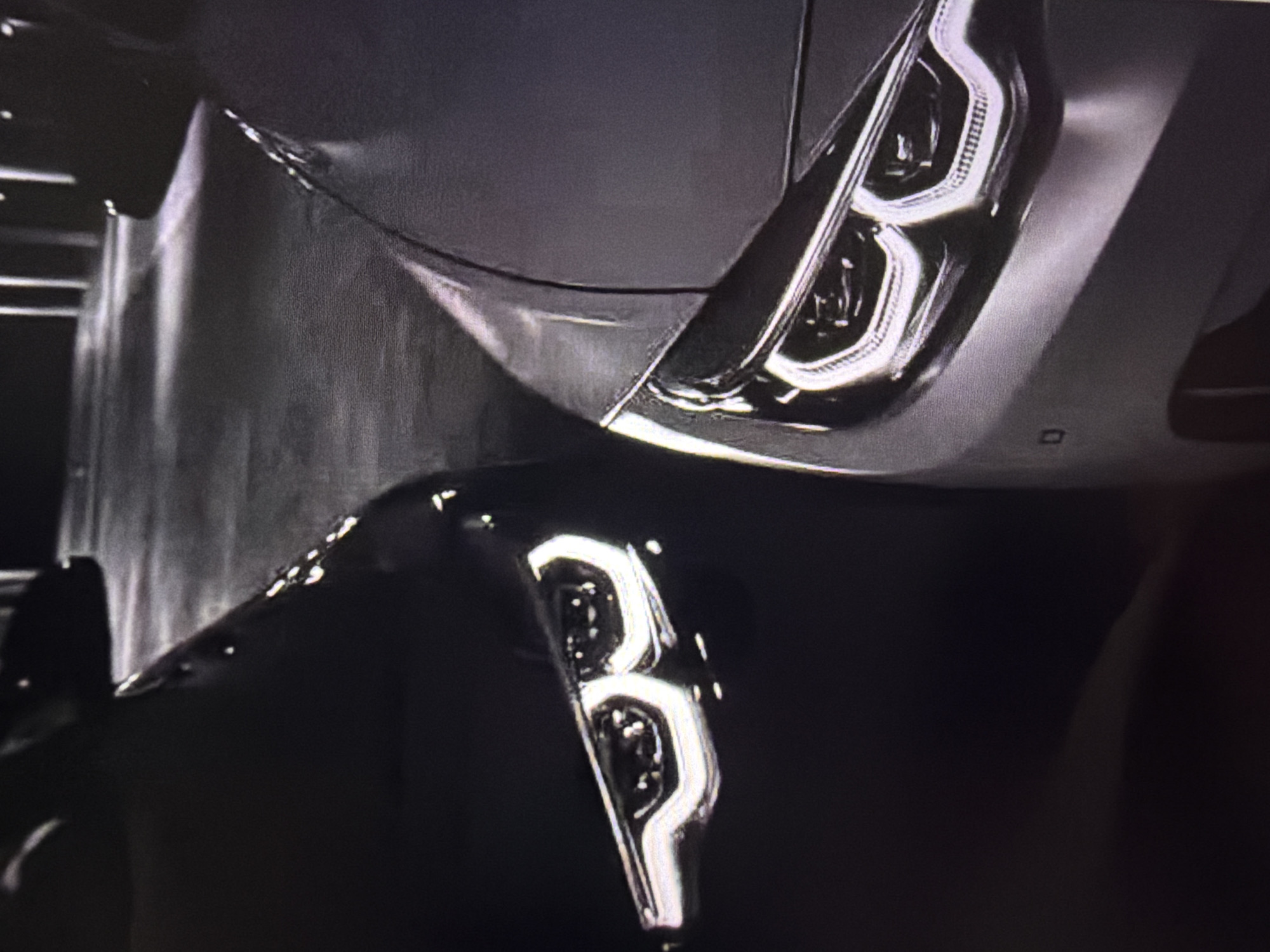 The image shows the front headlights of two cars parked side-by-side. The car on the left is dark, possibly black, and its headlight features a distinctive "angel eye" or halo-like daytime running light. The car on the right appears to be lighter in color, possibly silver or grey, and also has a similar "angel eye" headlight design. Both headlights are illuminated. The background is dark and appears to be an urban or industrial setting, with vertical structures visible in the distance, possibly lampposts or pillars. The ground surface seems to be paved and reflective. The image is a close-up, focusing on the front ends of the vehicles. The image does not contain any discernible elements that would allow for the specific identification of the city of Beșalma, Moldova.