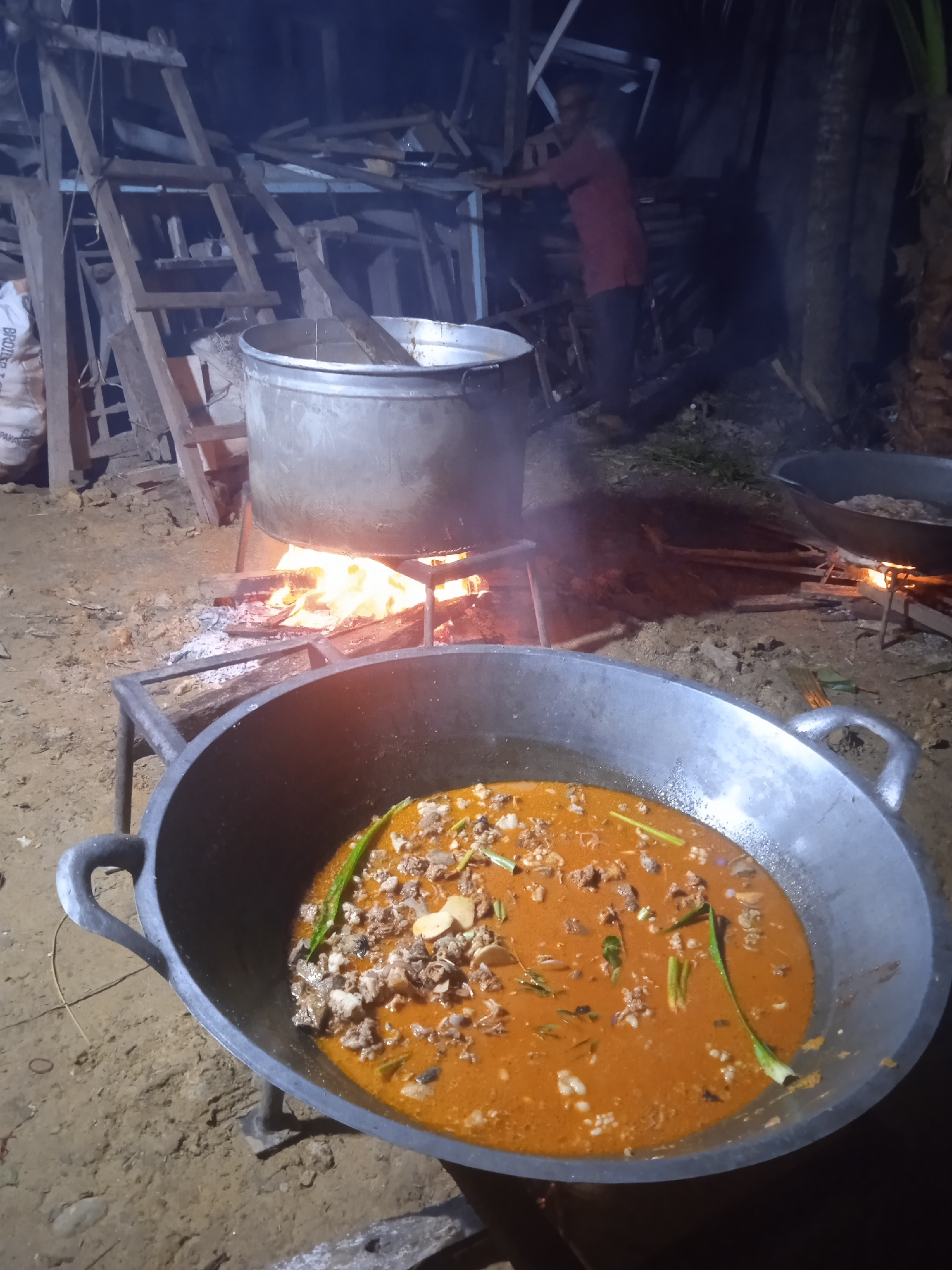 In the city of Keudeteupinpunti, Indonesia, a culinary scene unfolds at dusk. A large, weathered wok, filled with a rich, reddish-orange stew, sits in the foreground. The stew contains visible pieces of meat, green herbs, and what appear to be small shells or mollusks. The wok is supported by a crude metal stand over a dirt ground.
Behind the wok, a large pot, also on a metal stand, is heated by a roaring fire of logs and embers. Flames lick up the sides of the pot, casting a warm glow. In the background, a person dressed in a red shirt and dark pants stands near a pile of wooden structures and lumber, possibly a makeshift shelter or storage area. A ladder leans against some of the wooden debris. The overall environment appears to be outdoors, with the ground covered in dirt and scattered vegetation. The lighting suggests it is either early evening or late night.