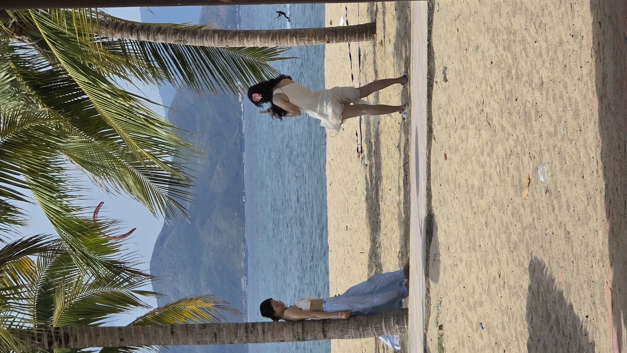 The image depicts a coastal scene, likely in Nha Trang Ward, Vietnam, showing two women interacting with palm trees on a beach. One woman is lying on a fallen palm tree trunk, wearing a white top and a blue and white checkered skirt. The other woman is standing further down the beach, wearing a white dress and a flower in her hair. She seems to be taking a photo or video with her phone. The background features a blue sea with mountains in the distance, and the beach is covered with sand. Palm trees are prominent in the foreground and on the side of the beach.