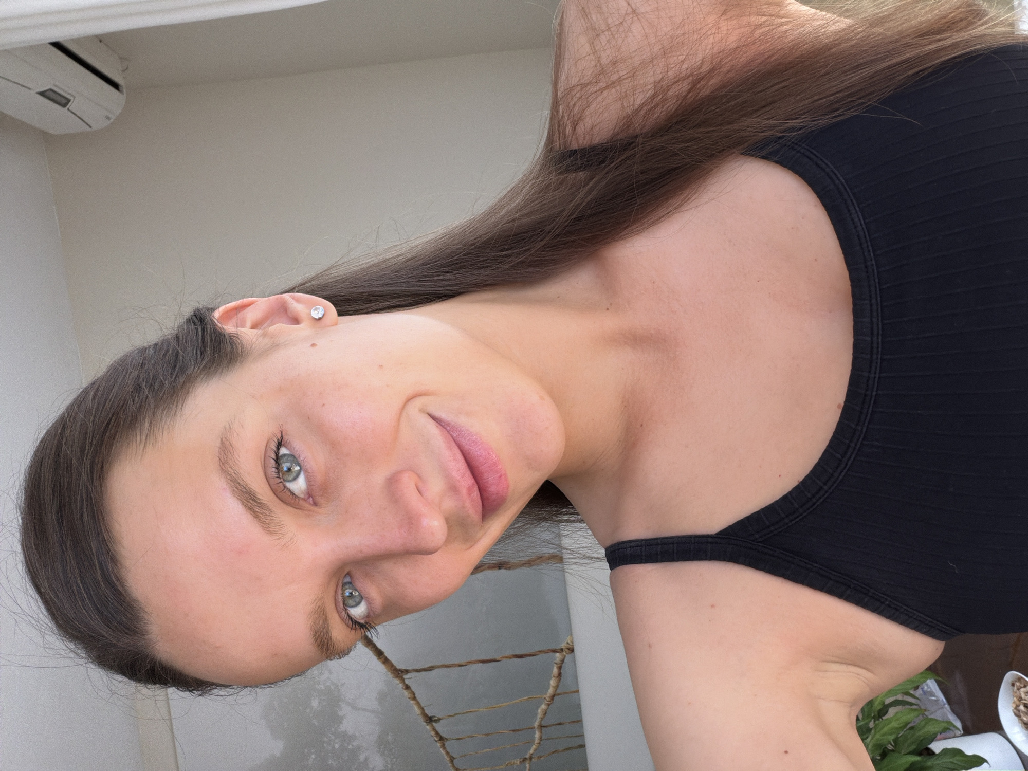 A young woman with long brown hair, wearing a black tank top, is looking at the camera. She has fair skin and light-colored eyes. The background includes a white wall with an air conditioning unit visible at the top left. Faintly visible behind the woman is a grey wall and a plant is visible in the bottom right corner of the frame. The location appears to be indoors, possibly a balcony of a building in Budva, Montenegro.