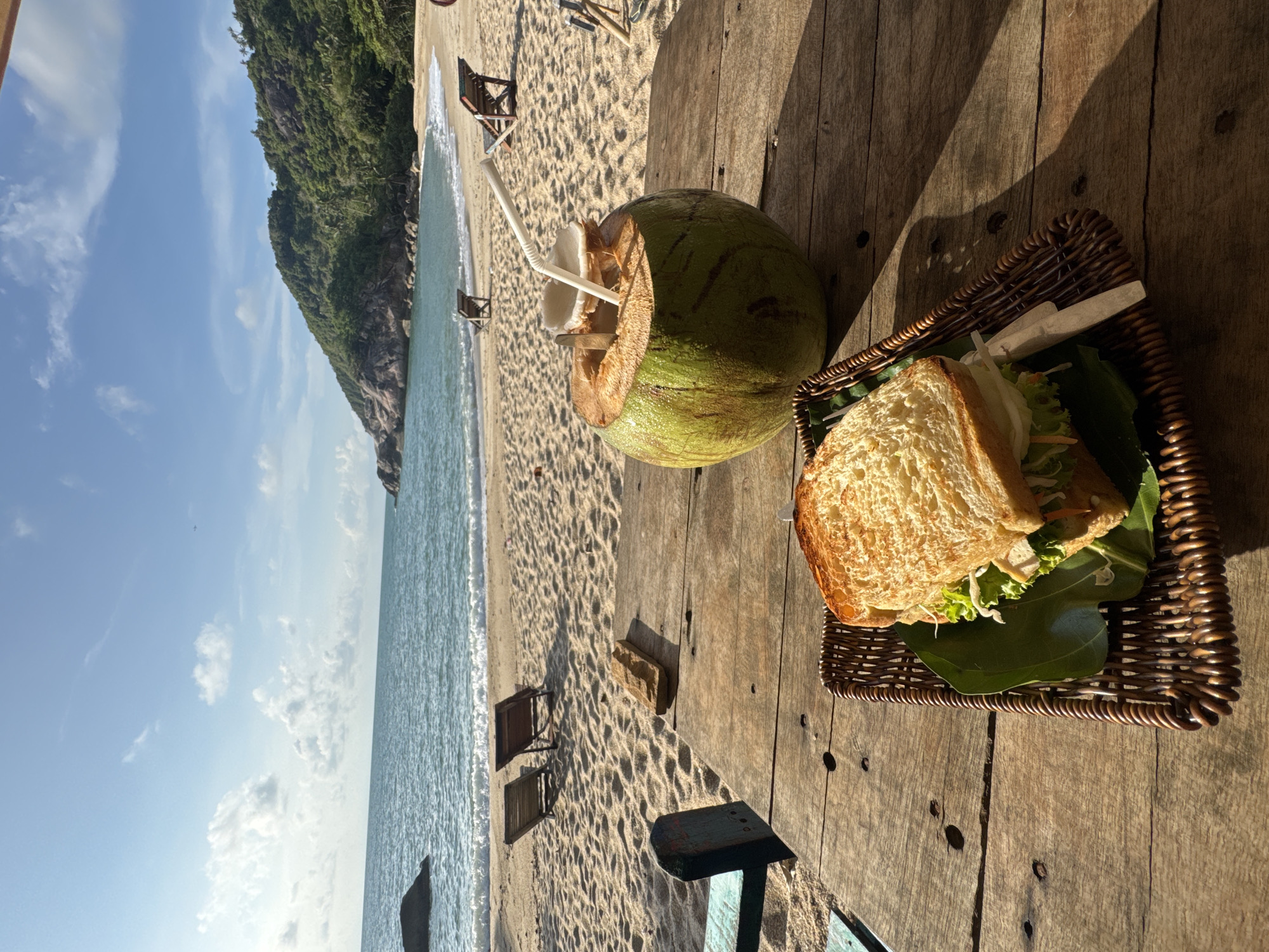 An eye-level, outdoor shot shows a tropical beach scene with a wooden table prominently featuring a coconut drink and a sandwich. The location is Baan Thong Nang, Thailand, as referenced in the prompt.
The right side of the shot focuses on the wooden table, where a partially cut green coconut with two white straws sits next to a woven basket containing a sandwich. The sandwich consists of toast, lettuce, and other fillings.
The left side of the shot reveals a beach with light brown sand, turquoise water, and a partially visible green, tree-covered hillside in the upper left corner. There are a couple of unoccupied beach chairs on the sand. The sky is light blue with scattered white clouds. The scene is lit by natural sunlight, casting long shadows on the wooden table.