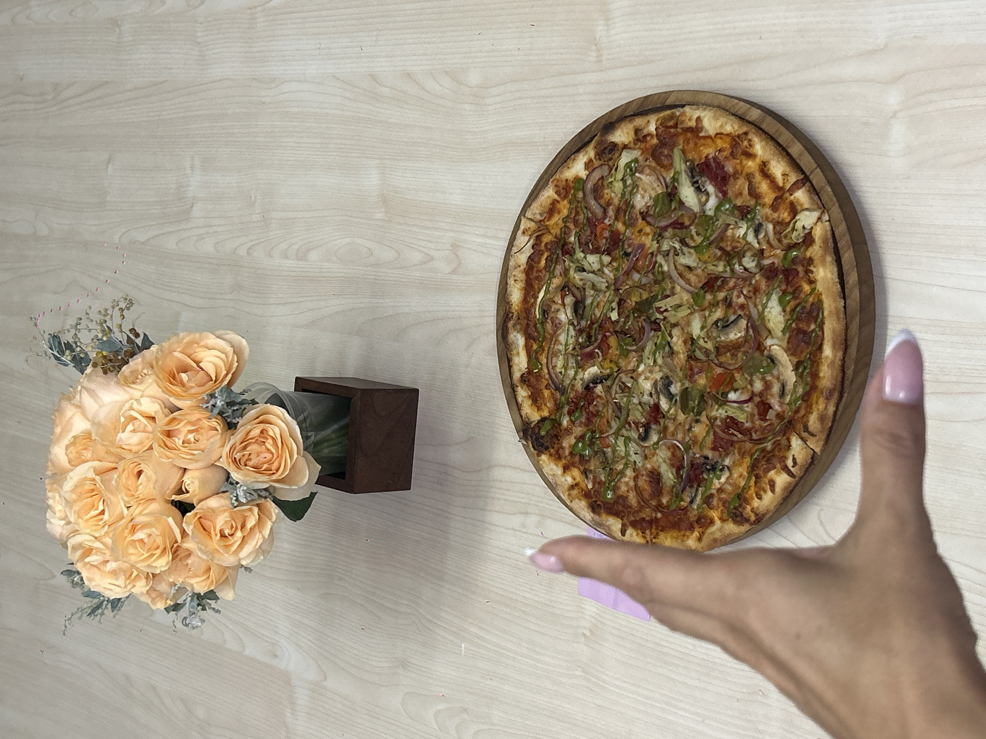 A overhead shot shows a pizza on a wooden board, next to a bouquet of peach roses in a dark brown wooden vase. A hand with pink nail polish is reaching towards the pizza. The scene is set on a light-colored wooden table. This image was taken in Kuta Selatan, Indonesia.