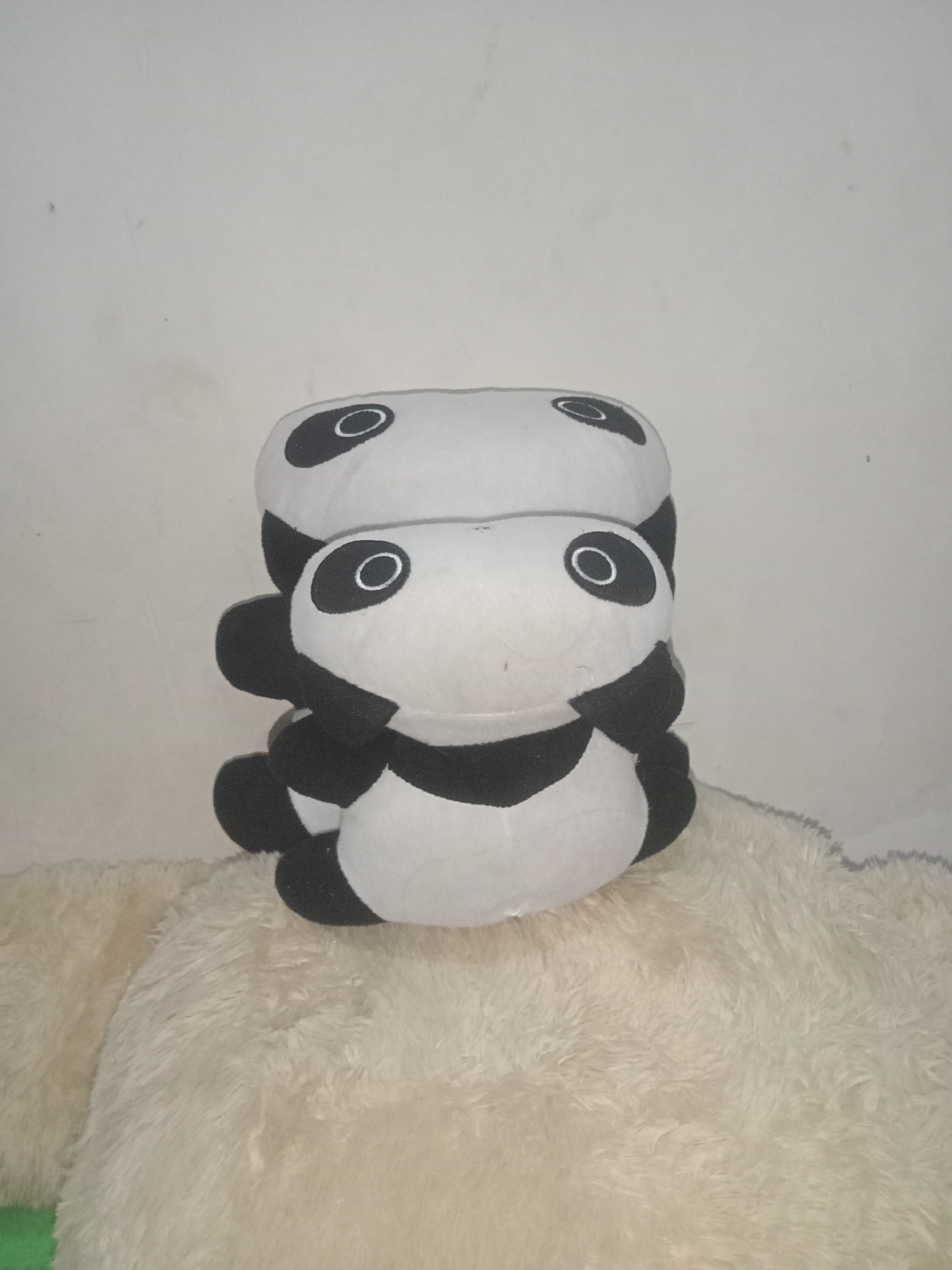 The image shows a stack of three plush panda toys. The pandas are black and white with distinctive eye patches. They are positioned on a fluffy, cream-colored surface. The background is a plain, light-colored wall with some subtle texture and marks. There is no visible indication of the location being the city of Tanggaran, Indonesia. The scene is static and focuses solely on the stuffed toys.