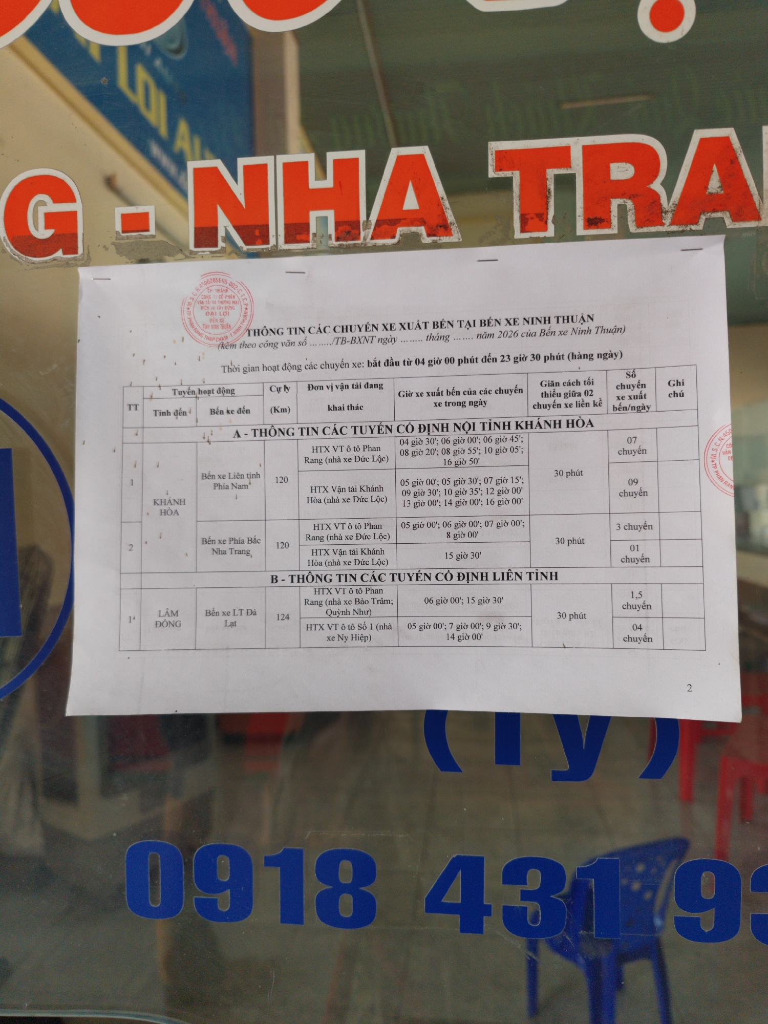 The image shows a document posted on a window, providing information about bus routes departing from Ninh Thuan Bus Station in Phan Rang – Tháp Chàm, Vietnam. The document, titled "Information on Bus Routes Departing from Ninh Thuan Bus Station," is dated 2026. It details routes to Khanh Hoa province (both north and south) and Lam Dong province, specifically to Da Lat. The schedule includes departure times, distance in kilometers, transportation operators, time intervals between buses, and the number of daily departures. The window has a partial view of the words "G-NHA TRA" in red and blue, and a phone number, 0918 43193, is visible at the bottom. Some chairs are seen through the glass.