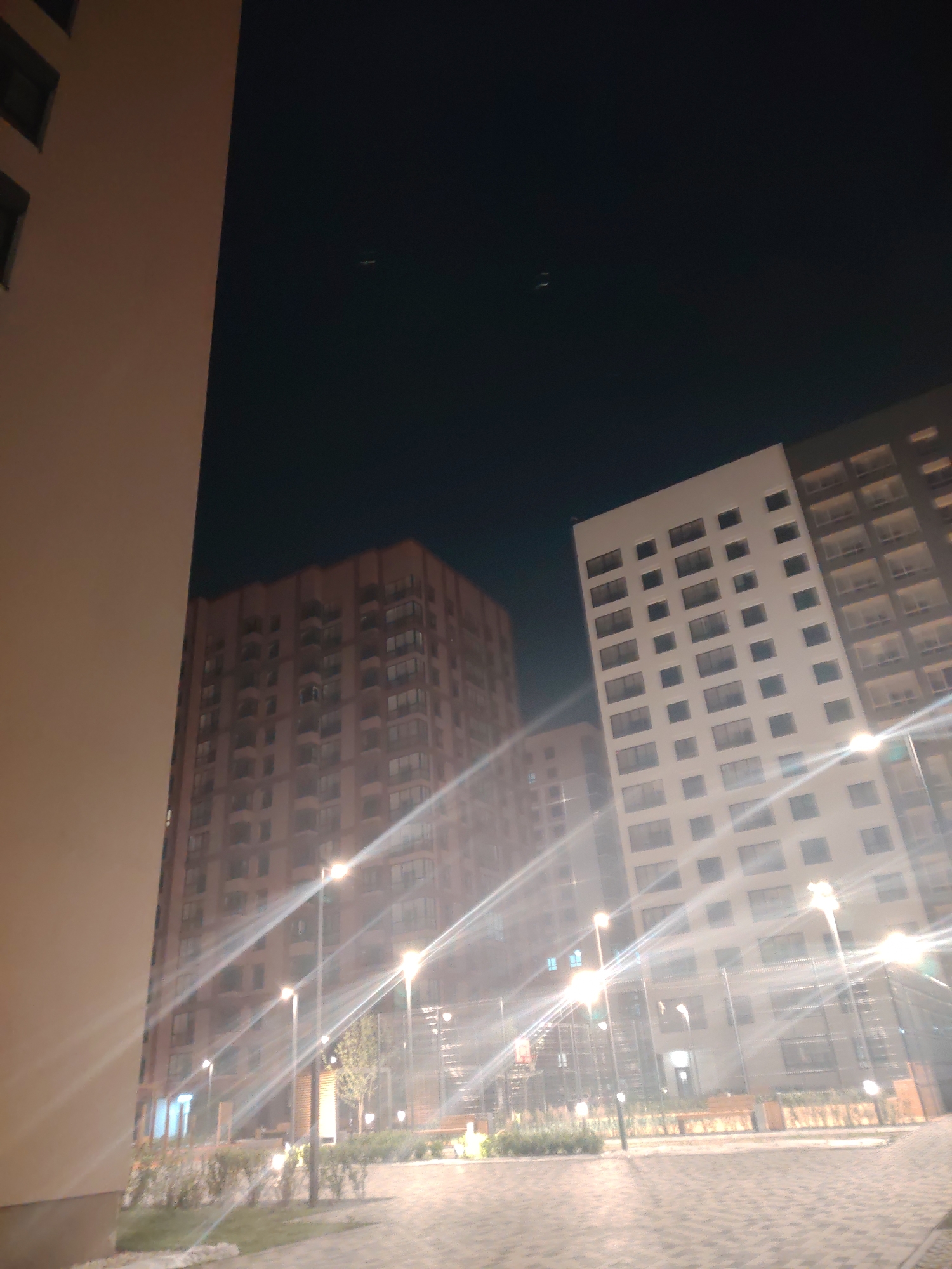 The image depicts an outdoor scene at night, focusing on modern residential buildings. Two tall buildings are prominent: one is a lighter-colored, modern structure with a grid of windows, and the other is a taller, reddish-brown building with a more intricate facade and numerous balconies. Between them, a shorter, lighter-colored building is visible. The foreground shows a paved walkway with streetlights casting a bright, starburst effect on the image due to the long exposure. There are also areas of landscaping with low bushes and trees. The sky is dark, indicating nighttime, with two faint specks that could be distant aircraft. The lighting creates a somewhat surreal and atmospheric mood. No people are visible, and no text is present.