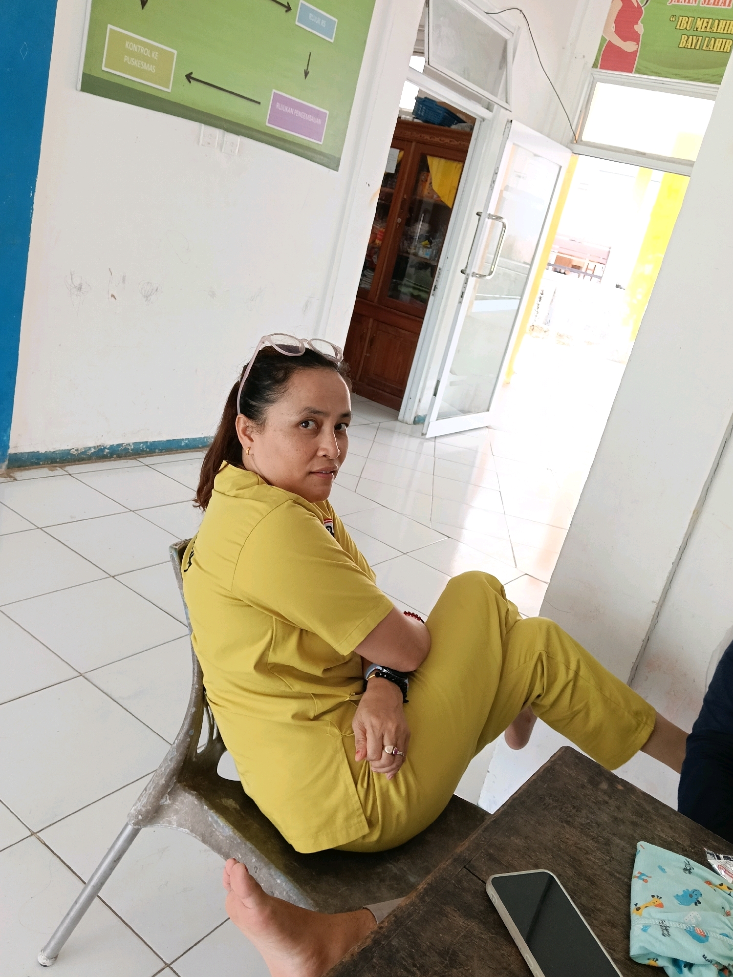 In a brightly lit room with white tiled floors, a woman in a yellow uniform sits on a metallic chair. She has her glasses perched on top of her head and is looking over her right shoulder at the camera. Her posture suggests she is relaxed, with her legs crossed. A wooden table is in front of her, displaying a smartphone and a folded piece of patterned fabric. In the background, a door stands ajar, revealing a glimpse of another room. To the left, a green poster with a diagram is affixed to the wall. The scene appears to be within a healthcare facility in Muntei, Indonesia.