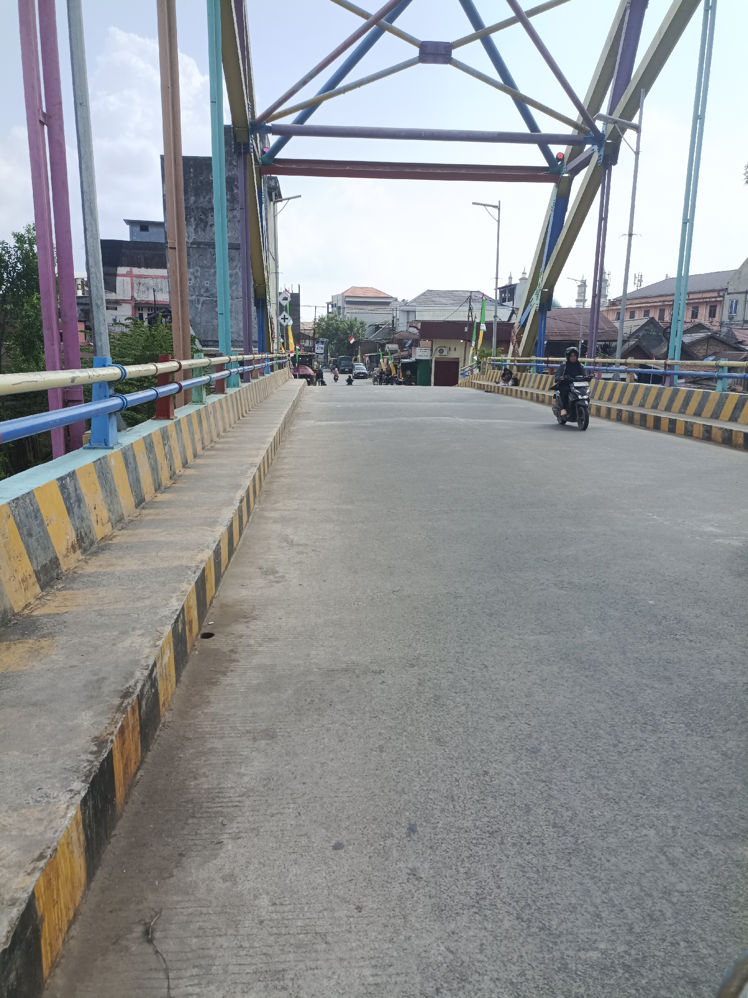 The image depicts a view from a bridge in Sangatta, Indonesia. The bridge features a colorful metal superstructure with beams painted in yellow, blue, and purple. The railings are painted in sections of blue and yellow with black stripes. The road surface is asphalt with distinct yellow and black striped concrete barriers along the edge. In the distance, buildings with various architectural styles are visible. Several motorbikes are present on the road, with one rider in the foreground wearing a dark outfit and headscarf. The sky is bright and overcast.
