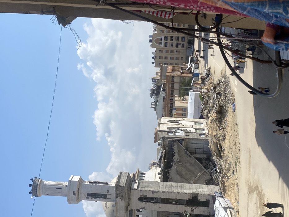 An elevated view captures a street scene in Khan Yunis, Palestinian Territory, under a bright blue sky with scattered white cumulus clouds. On the far left, a mosque features a tall, cylindrical minaret, largely intact but showing signs of wear and damage at its base, topped with multiple loudspeakers. The main structure of the mosque adjacent to the minaret is severely damaged, with large sections of its facade and interior walls collapsed and exposed.
To the right of the mosque, a multi-story building is extensively destroyed, with a significant portion of its upper floors and interior completely collapsed, forming a large pile of concrete rubble, twisted rebar, and debris at ground level that extends across a dirt and sand area. A white tent structure is visible near the base of the damaged buildings on the left.
The foreground and mid-ground feature a paved road, partially obscured by shadows and further piles of rubble and sand. Numerous individuals, appearing to be civilians, including adults and children, are walking on the road. A faint pedestrian crossing is visible.
On the far right, the image is framed by a partial view of an adjacent building structure, possibly a balcony, with exposed rebar and electrical wiring extending from a damaged concrete edge. Draped fabric, patterned in red, blue, and white, hangs over a railing in the immediate foreground, partially obscuring the lower right corner.
In the mid-distance and background, several multi-story residential and commercial buildings are visible. Some exhibit apparent damage, including darkened areas on rooftops, collapsed sections, and exposed interiors. Numerous black water tanks are visible on the rooftops of some buildings. Overhead electrical wires traverse the scene against the sky.
