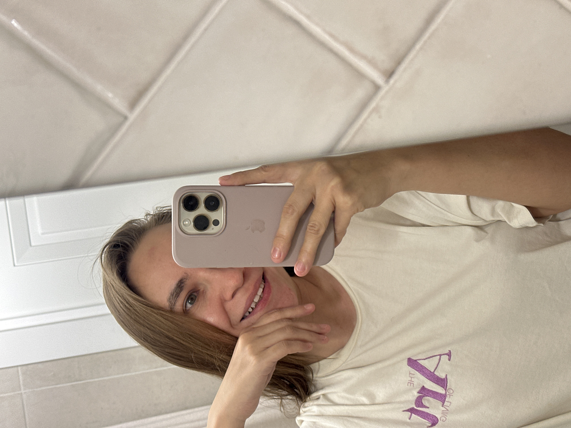 A woman is taking a selfie in a bathroom. She is holding a lilac iPhone in front of her face with her right hand, partially obscuring her forehead and right eye. Her left hand is near her chin. She is smiling, exposing her teeth. She has light brown hair and green eyes. She is wearing a light beige t-shirt with "The Art of Living" printed in purple. The bathroom has white tiled walls. The image offers no indication of Kharnavy, Russia.