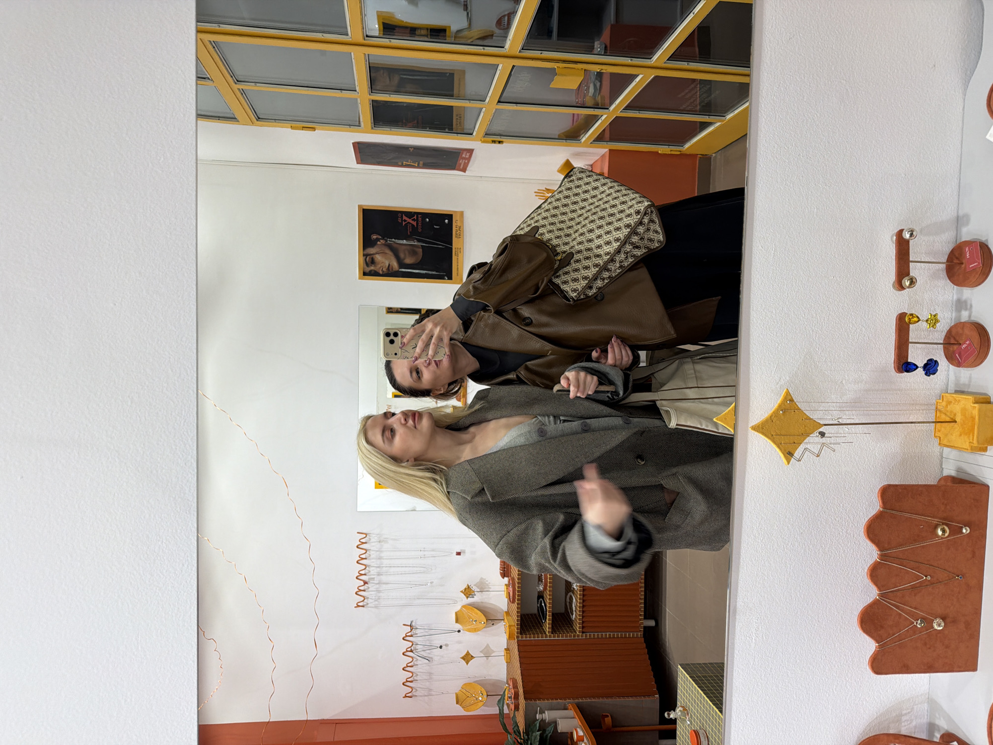 The image captures a mirror selfie taken by two women inside a contemporary retail space, likely a jewelry or accessories boutique in Belgrade, Serbia.
In the reflection, on the left, a blonde woman is visible, wearing a gray herringbone-patterned coat over a gray top. She is posing casually, looking slightly upwards, with her right hand slightly blurred from motion. To her right and slightly behind her, a second woman with darker hair holds a smartphone up to capture their reflection. She is dressed in a brown leather-like jacket over a dark top and carries a distinctive beige and brown patterned bag over her shoulder. Her face is visible in the phone's screen reflection.
The surrounding environment, seen in the mirror, is a shop interior. Various jewelry displays are prominent. On the right side of the reflection, vertical stands hold small earrings, including designs resembling a gold star and a blue flower. Below these are rust-colored, wavy-edged velvet stands showcasing necklaces and pendants. Similar velvet displays and abstract yellow star or diamond-shaped displays with hanging metallic pieces are visible on the left side of the reflection.
The background shows yellow-framed glass partitions or windows at the top. Below these, posters are affixed to a white wall; one poster features a person's profile and displays the text "ANGELO X LE VIE," while another is a solid reddish-orange. Further down, there are wooden or orange-toned display cabinets and shelving, with one section featuring a grid pattern. A small potted plant is also visible on a lower shelf. Small tags on some jewelry items or displays bear the text "PROSI." The setting is brightly lit, suggesting an indoor daytime scene within a modern store featuring a warm color palette.