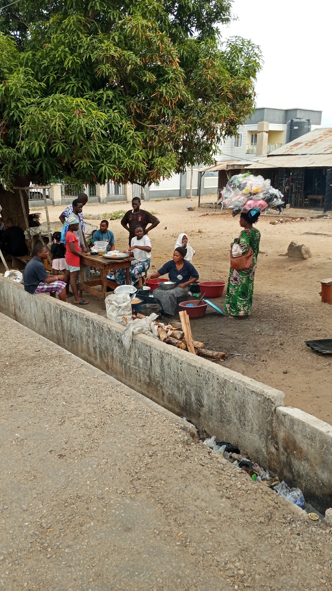 The image shows an outdoor scene in Zango-ndaji, Nigeria. A group of people is gathered around a makeshift cooking area located next to a concrete drainage ditch. A woman is actively cooking food over a wood fire, using a large pot. Several other people are seated or standing nearby, some gathered around a wooden table where food is being served or eaten. One woman stands on the right side of the frame, wearing a green dress and carrying a handbag. Behind the group, there are buildings of varying construction types, including a simple structure with a corrugated metal roof and a more modern building in the background. The area appears to be a public space with a dirt surface. The drainage ditch is filled with trash and debris. A large tree provides shade over the cooking area.