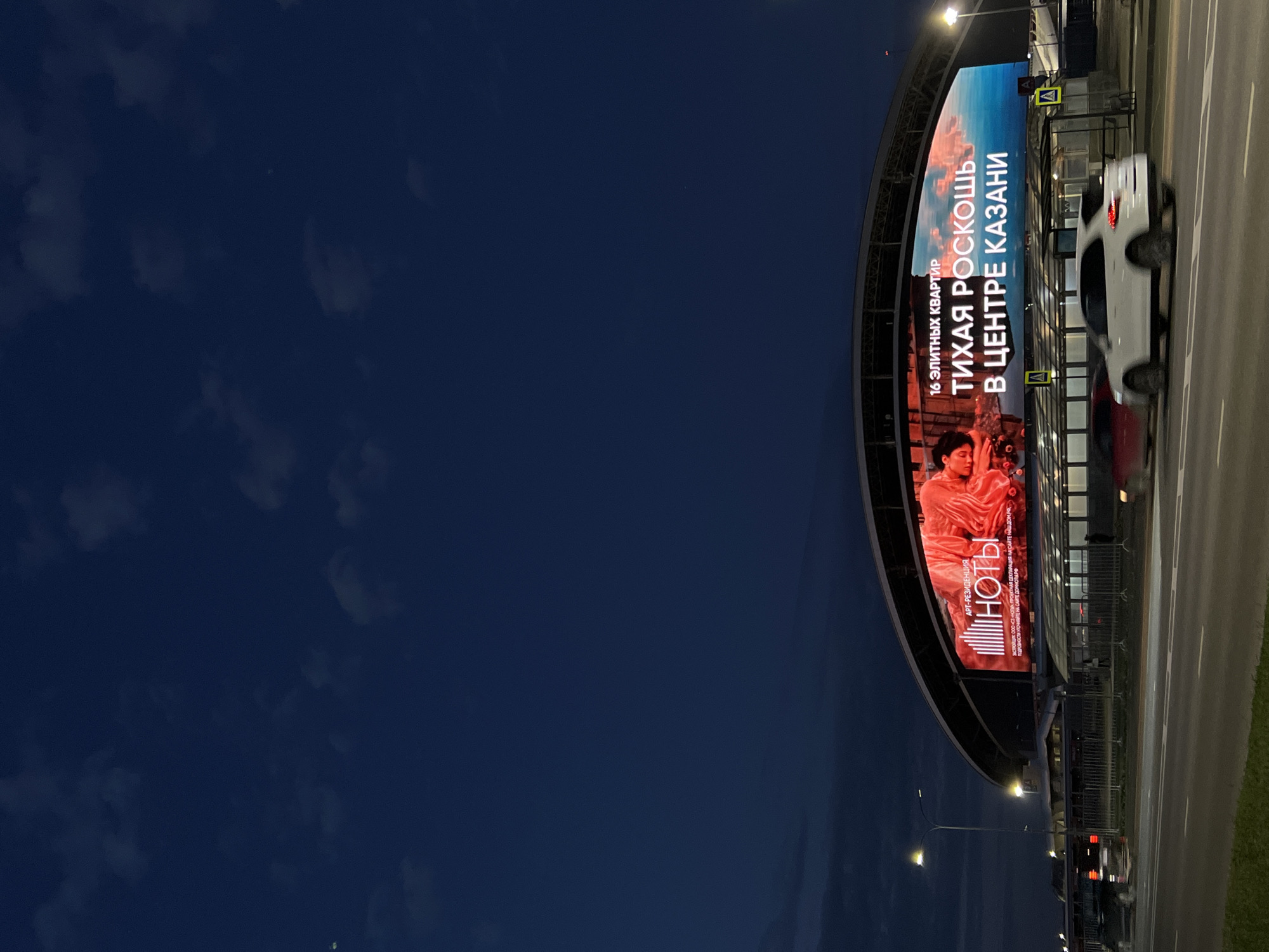 The image shows a large LED billboard displaying an advertisement at night in Kazan, Russia. The billboard is situated on the exterior of a modern, curved building, likely a stadium or exhibition center, with a large, arched roof structure. The advertisement features text in Russian, with the primary message reading "ТИХАЯ РОСКОШЬ В ЦЕНТРЕ КАЗАНИ" (Quiet Luxury in the Center of Kazan). Above this, it states "16 ЭЛИТНЫХ КВАРТИР" (16 Elite Apartments). There is also a logo for "НОТЫ" and a smaller line of text. The advertisement includes a visual element on the left side, depicting a person in red fabric against a red and white abstract background.
The scene is set at dusk or night, with a dark blue sky and scattered clouds. Streetlights are visible, illuminating the area. In the foreground, a road with white lane markings is present, with two cars in motion. One car is red and appears to be moving away from the viewer, while a white SUV is in the right lane, also moving away. A pedestrian walkway with a glass enclosure and yellow warning signs is visible beneath the billboard. The overall environment suggests an urban setting, likely a developed area with transportation infrastructure and commercial or event facilities.