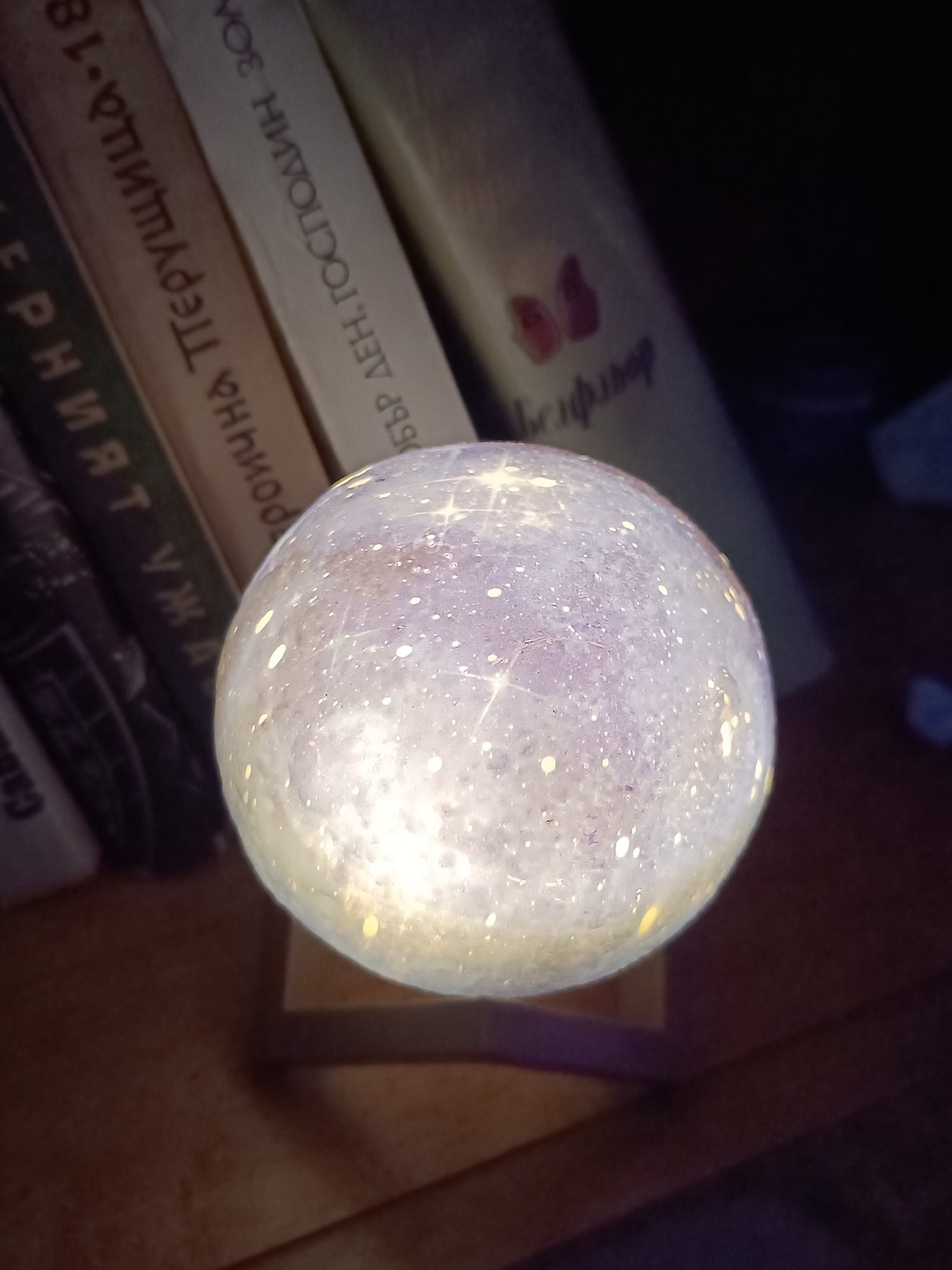 A close-up shot captures a glowing spherical object, likely a decorative lamp, resting on a wooden stand. The lamp emits a bright, diffused light with a pattern reminiscent of stars or planetary bodies. It is situated against a backdrop of several books with spines facing the viewer. The book spines display Cyrillic text. The location information suggests this photo was taken in Cherkovitsa, Bulgaria.