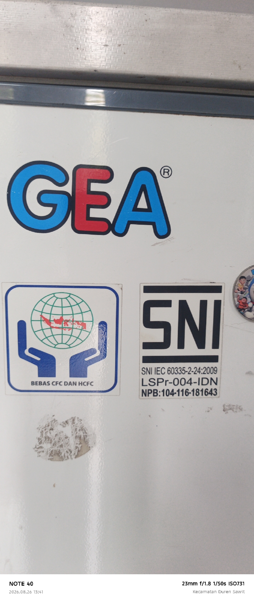 The image shows a close-up of a white surface, likely a refrigerator, with several stickers and branding. Dominating the upper half of the image is the blue and red "GEA" logo with a registered trademark symbol. Below the GEA logo, on the left, is a square sticker with a blue border containing a graphic of two blue hands holding a globe with red outlines of landmasses, and text that reads "BEBAS CFC DAN HCFC". To the right of this sticker is another rectangular sticker with a black border and bold black text reading "SNI", followed by "SNI IEC 60335-2-24:2009", "LSPpr-004-IDN", and "NPB:104-116-181643". A portion of another sticker with cartoon characters is visible on the far right. The bottom of the image contains text overlays: "NOTE 40", "2026.08.26 13:41", "23mm f/1.8 1/50s ISO731", and "Kecamatan Duren Sawit". "Kecamatan Duren Sawit" indicates the location is Duren Sawit District in East Jakarta, Indonesia. The surface shows some signs of wear and tear, with scuff marks and what appears to be peeling sticker residue.