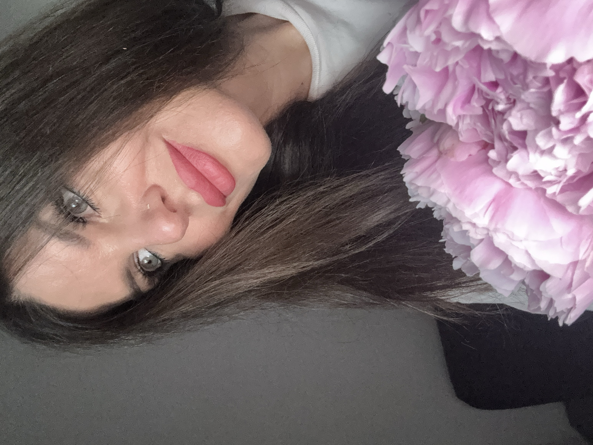 A close-up, head-and-shoulders shot captures a woman with long, dark brown hair and striking gray-blue eyes. Her head is tilted slightly, and she is looking upwards and to her left, with a subtle smile playing on her lips. She is wearing a light-colored top, and a bouquet of large, pink peonies is held in the foreground, obscuring the lower part of the image. The background is a plain, dark gray wall, suggesting an indoor setting. The image is taken from a slightly low angle. The woman has a nose ring and appears to be wearing makeup, including pink lipstick. The lighting suggests natural light, possibly from a window. There is no discernible connection to Chaikovsky, Russia, in the visual information provided.