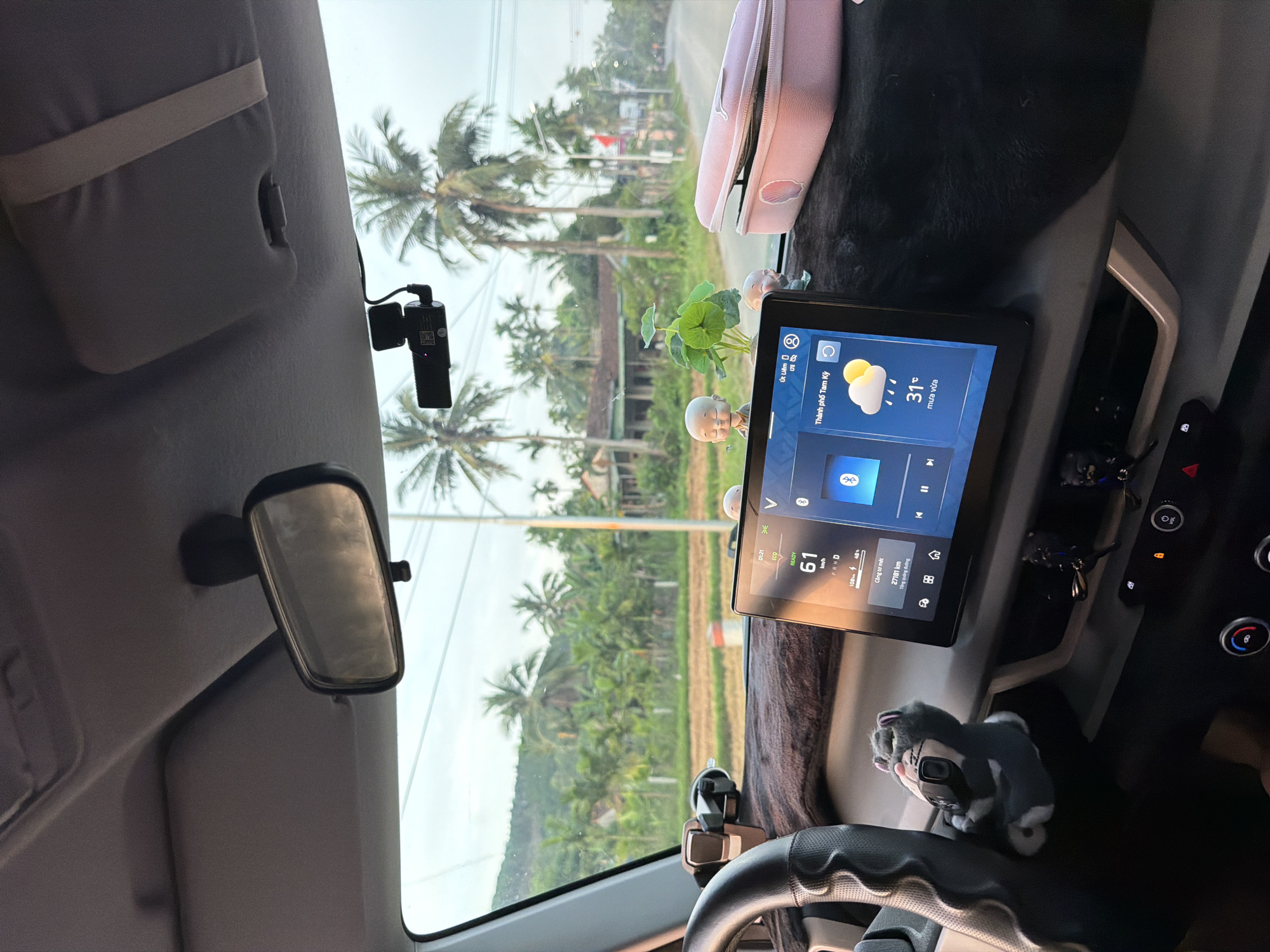 The image displays the interior of a car, viewed from the driver's perspective, looking out through the windshield. The car appears to be driving on a road bordered by lush tropical vegetation, featuring numerous tall palm trees. The bright sky indicates it is daytime.
A large digital display dominates the dashboard, showing various information. Its vehicle status section indicates "READY", a speed of "61 km/h", gear selection "P R N D", and power levels "108V 48%". The odometer reads "27181 km". A weather widget on the screen displays "Thành phố Tam Kỳ" (Tam Kỳ City), a temperature of "31°C", and "mưa vừa" (light rain). Other text visible on the screen includes "01:21 ECO" and "Út Liêm". A "V" logo, likely belonging to the car manufacturer VinFast, is present on the screen.
In the car's interior, a dashcam is mounted near the top of the windshield, and a rearview mirror is visible on the left. The dashboard is adorned with two small decorative figurines, a small plant with lotus-like leaves, and a pink pouch or pencil case. A grey and white stuffed animal rests near the steering wheel, and keys hang below the digital display. The tropical setting and the Vietnamese text confirm the location as Vietnam, specifically Tam Kỳ City, which aligns with the provided context of Bình Chương, Vietnam.
