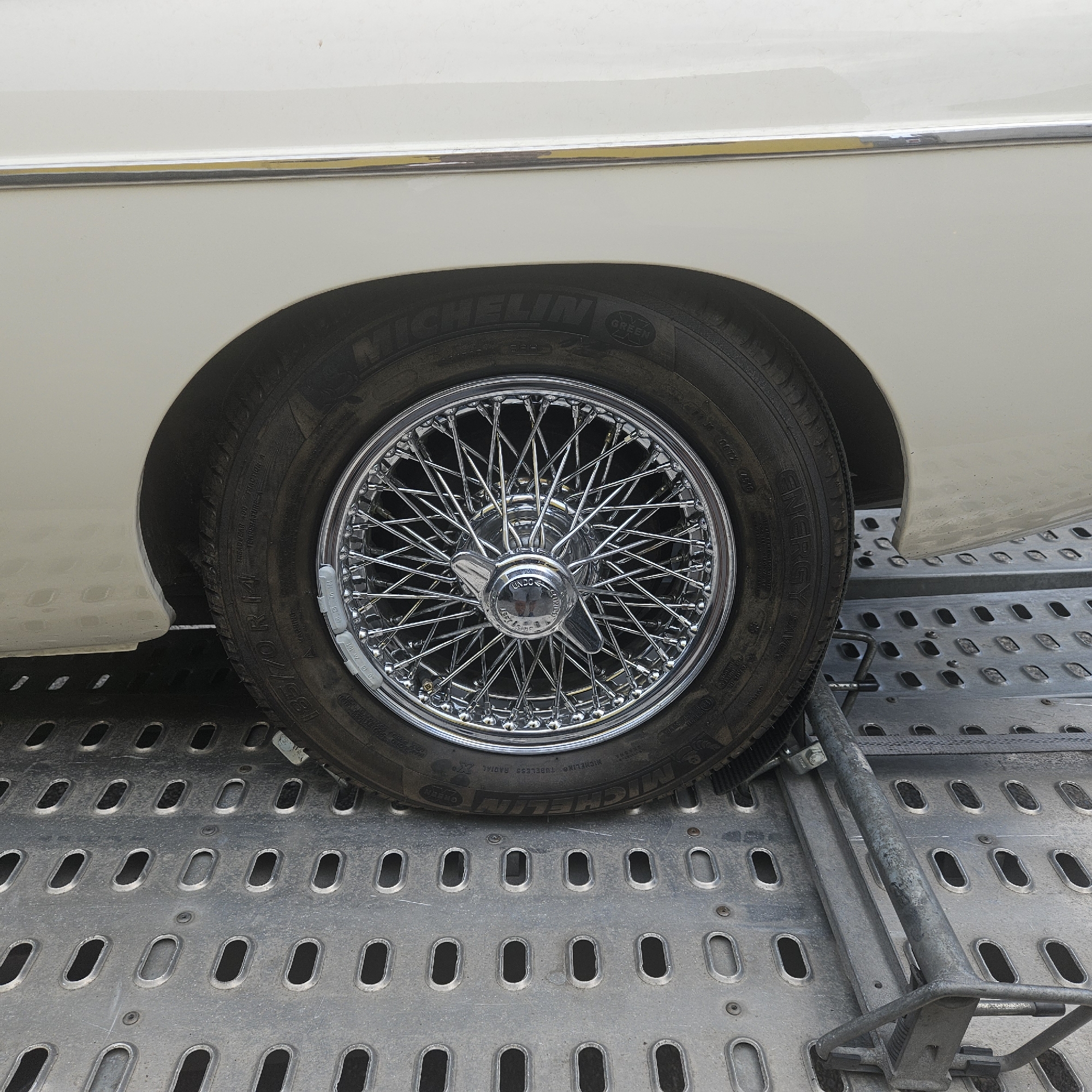 The image shows a close-up, high-angle view of a vintage car's wheel and lower body, likely parked on a metal trailer or loading dock. The car is a light color, possibly cream or off-white, with a chrome trim strip visible along the body panel above the wheel well. The wheel itself is a classic chrome wire-spoke design with a central hubcap. The tire is a Michelin radial, with visible markings including "185/70R14". There are small, grey wheel weights attached to the rim. The metal surface below the car is a gray, perforated platform with an industrial appearance, possibly part of a vehicle transport trailer. There are no people or animals visible in the frame. The scene is likely in a utilitarian or industrial area, consistent with a car transport or storage facility. The location of La Jonquera, Spain, is mentioned as a potential context for the image.