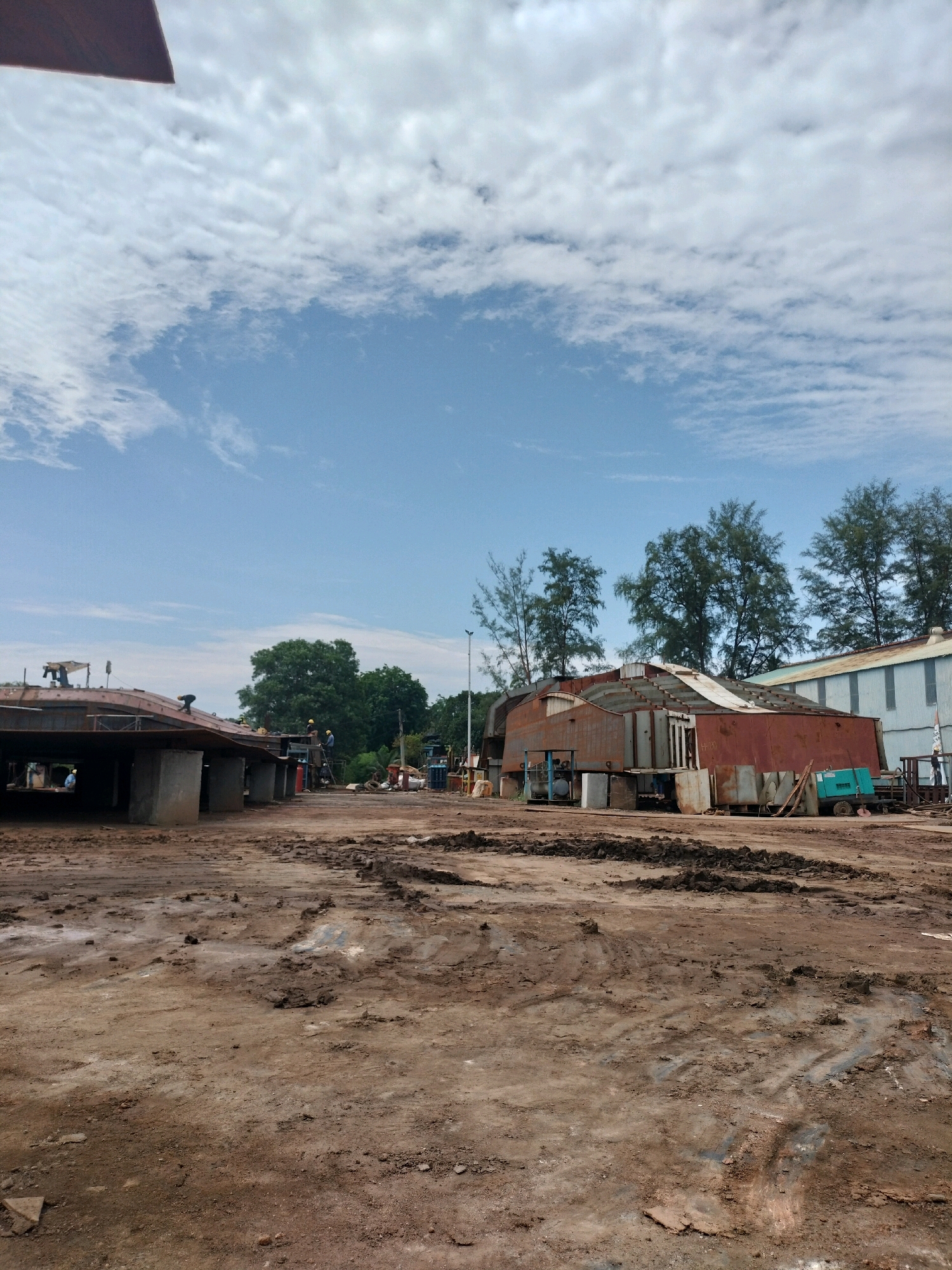 An outdoor, wide-angle, eye-level shot shows a construction or repair site with a large, unfinished metal structure in the foreground. The ground is covered in brown mud and dirt, with tire tracks visible. In the midground, several people in hard hats and safety vests are working on or near the metal structure, which appears to be part of a ship or boat hull. To the right, a green generator and other equipment are visible. In the background, several trees and buildings, including a long, corrugated metal building, are present. The sky is blue with white, wispy clouds. This scene is consistent with an industrial shipyard in Batam, Indonesia, which is known for its shipbuilding and repair facilities.