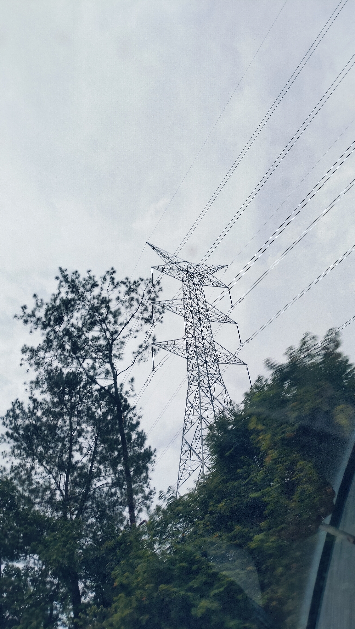 The image presents an outdoor scene captured from the perspective of someone inside a moving vehicle. A prominent feature is a tall, metallic electricity pylon (power tower) positioned slightly to the right of the center, its intricate lattice structure extending upwards towards the sky. Several power lines emanate from the pylon, stretching diagonally across the top right portion of the frame.
Dense green trees frame the scene on both sides. On the left, tall, dark green trees, possibly conifers, reach high into the frame, partially obscuring the pylon's base. On the right, a mass of green foliage appears somewhat blurred, indicating motion, and suggests the vehicle is traveling past. The bottom right corner of the image subtly reveals a car window frame or reflection, confirming the in-vehicle perspective.
The sky is uniformly overcast, a light grey without direct sunlight, suggesting a cloudy day or perhaps early morning/late afternoon. There are no visible people or specific activities taking place, only the observation of the power infrastructure and natural landscape from transit. No text is visible in the image. The setting is likely a rural or semi-urban area in Lamtamot, Indonesia, where such large pylons are part of the landscape. The overall mood is one of quiet observation of the environment during travel.