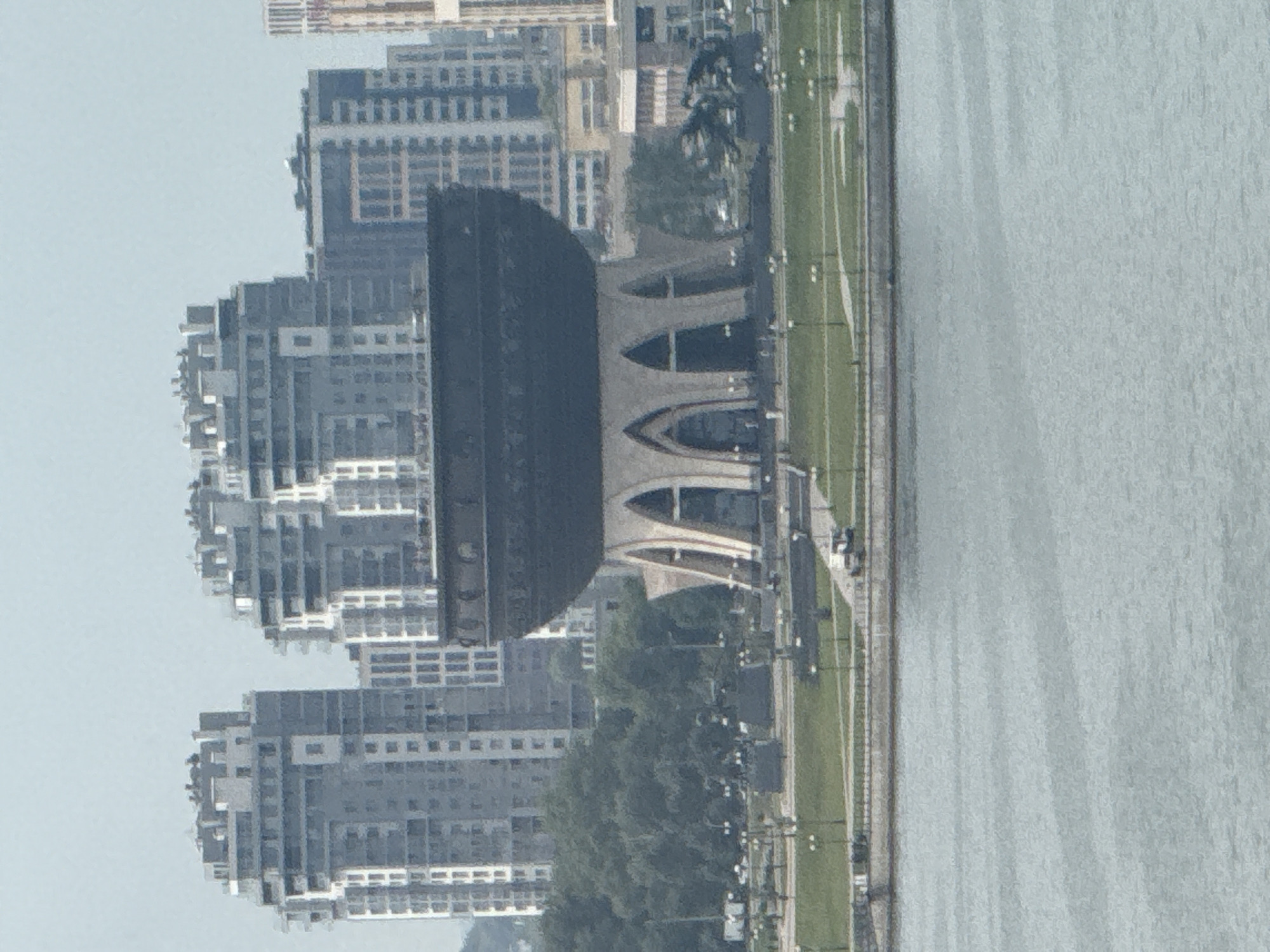 The image captures a wide shot of the Kazan Family Center, a distinctive building in Kazan, Russia, with the Kaban Lake in the foreground. The building, shaped like a cauldron, is a prominent feature on the embankment. In the background, modern high-rise apartment buildings are visible, suggesting an urban environment. The sky is overcast and pale, contributing to a muted color palette. The lake water is a calm blue-gray. The scene appears to be public, with park-like areas and pathways in front of the Family Center. There are no people clearly visible in the image, and the overall impression is one of civic architecture and urban development along a waterfront.