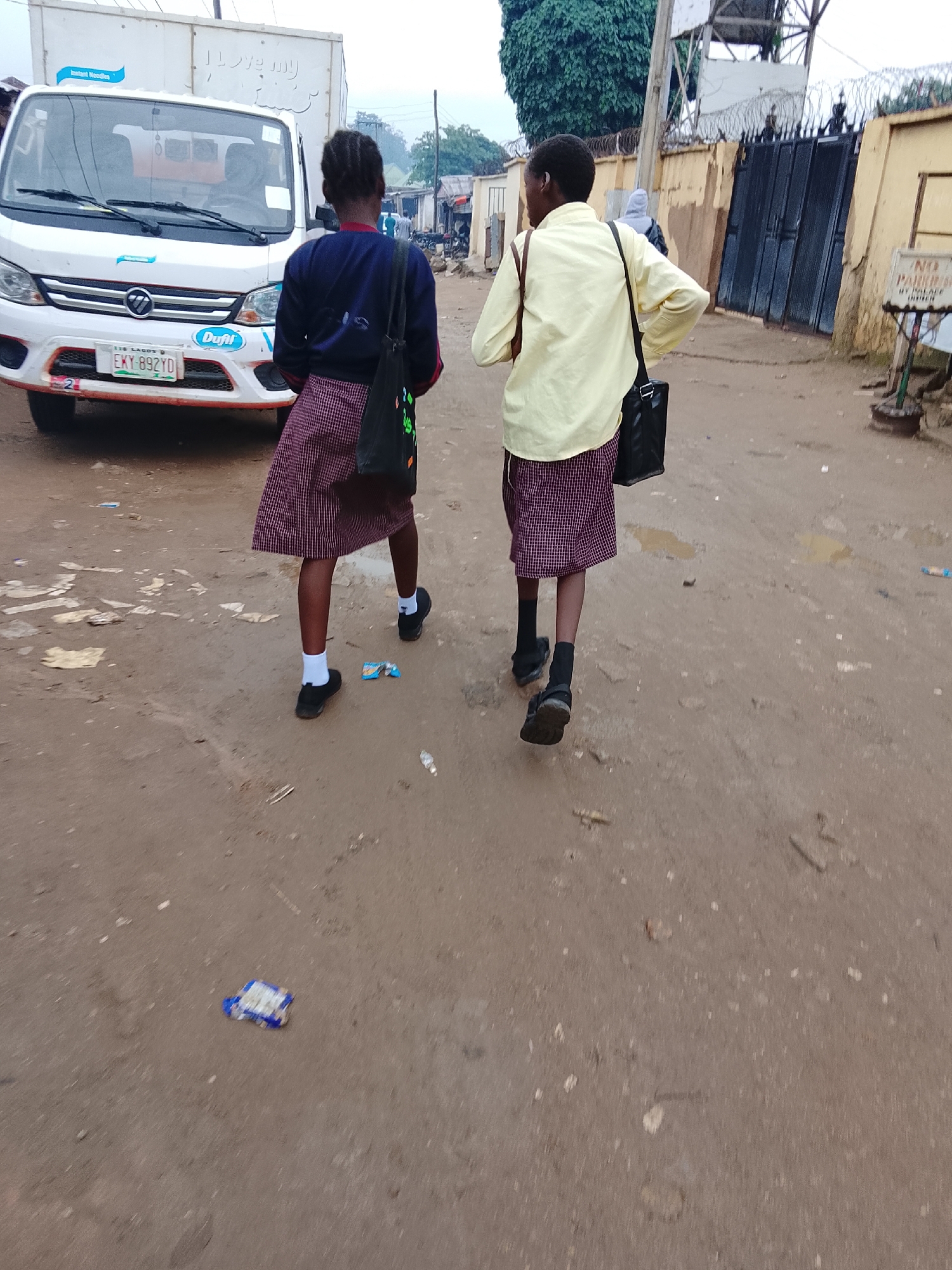 Two young girls, likely students, are walking away from the camera on a dirt road in Abuja, Nigeria. The girl on the left wears a dark blue long-sleeved shirt and a maroon and white checkered skirt, with white socks and black shoes. She carries a black bag with colorful patterns over her shoulder. The girl on the right wears a light yellow long-sleeved shirt and a similar maroon and white checkered skirt, with black socks and black shoes. She also carries a black bag over her shoulder.
In the background, a white delivery truck with "Instant Noodles" and "I love my family" branding is parked. Further down the road, there are buildings, a black metal gate with barbed wire, and a "NO PARKING" sign. The ground is muddy and littered with debris, suggesting a potentially unpaved or poorly maintained area. The sky appears overcast.