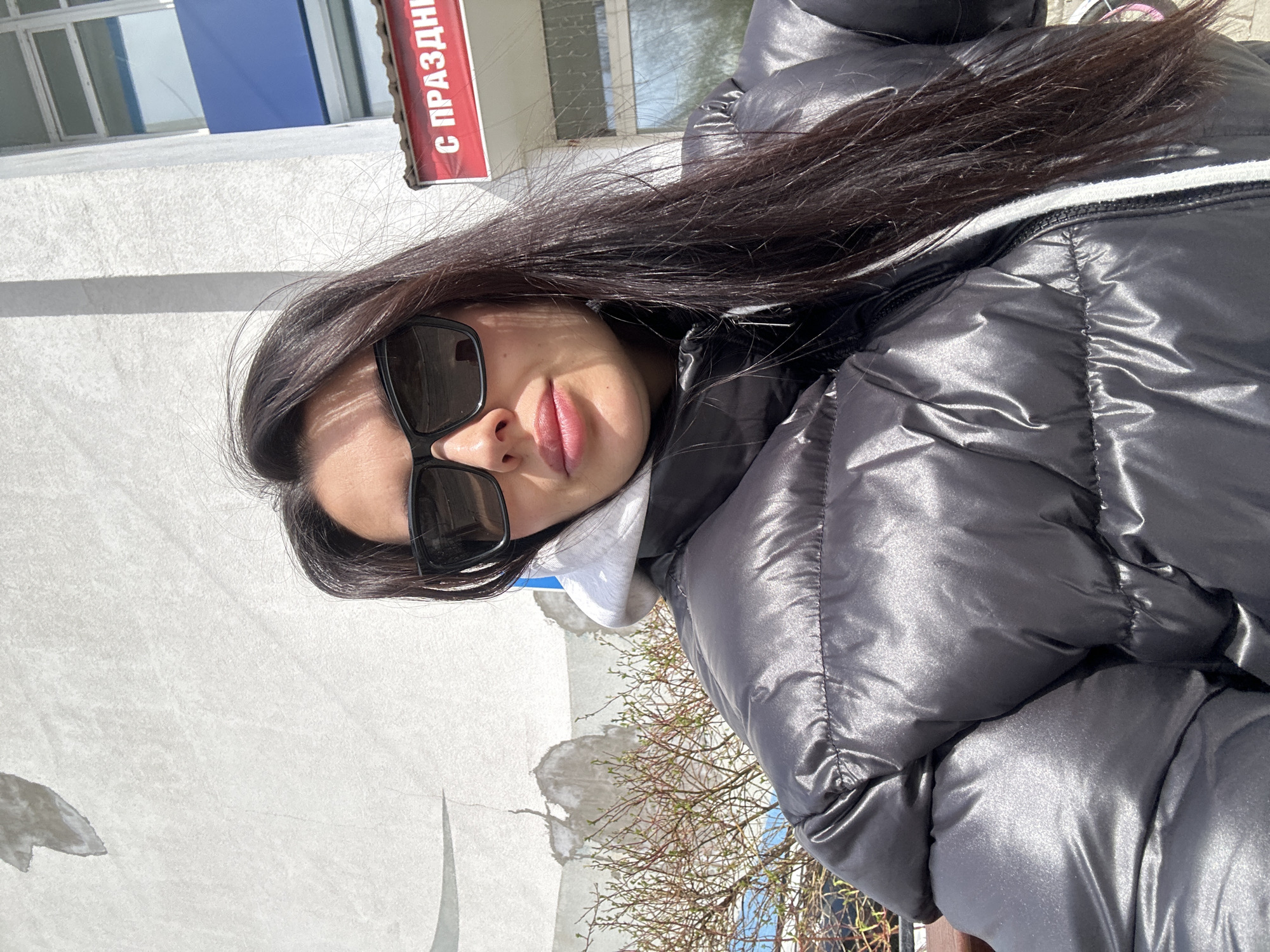A young woman is depicted in a close-up, selfie-style shot taken outdoors on a bright, sunny day. She has long, dark, straight hair and is wearing black sunglasses, which obscure her eyes. Her lips are slightly parted in a hint of a smile. She is dressed in a glossy, black puffer jacket, with the hood of a light-colored (possibly white or light grey) hoodie visible underneath.
The background features a light-colored, possibly white or grey, building with a textured surface. Above her head, a dark blue architectural element, possibly a window or awning, is visible on the building. To the upper right, a red sign is mounted on the building, displaying partial Cyrillic text that reads "С ПРАЗДНИ" (S PRAZDNI), likely part of "С ПРАЗДНИКОМ" (Happy Holiday). In the lower right of the image, bare tree branches with small green buds are visible, suggesting a springtime setting. The strong sunlight creates clear shadows and highlights the reflective quality of her jacket. The scene takes place in Chaikovsky, Russia.