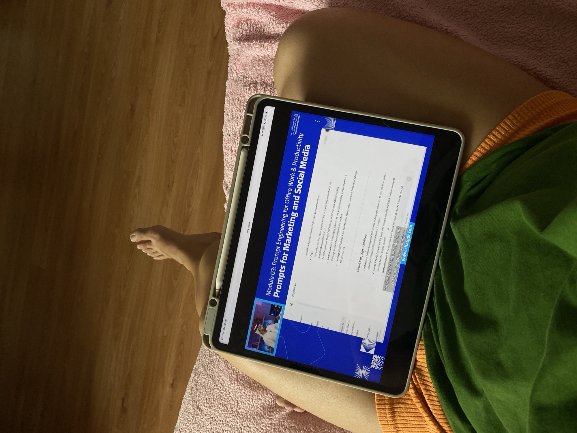 A person's legs are visible from the knee down, with one foot resting on a wooden floor. The person is lying down on a pink fuzzy surface, wearing an orange shirt and green shorts. They are holding a tablet computer, displaying a webpage with text and an image of a man in traditional Arab attire. The tablet screen also shows a timestamp of "3:01PM", network connectivity icons, and battery life indicator. The background is a wooden floor. The location of this scene is not specified within the provided image or its crops.