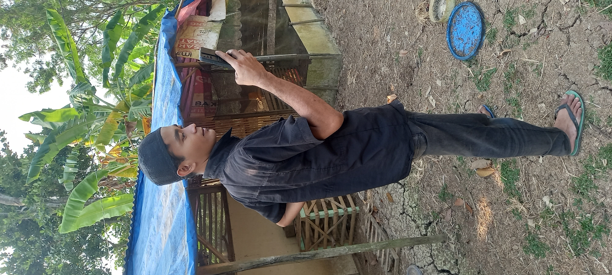 A young man wearing a black skullcap, black short-sleeved shirt, and dark grey jeans stands in an outdoor, rural environment in Sidamulya, Indonesia. He is holding a black smartphone in his right hand and looking at its screen. He is positioned facing to his right, with his left hand on his hip. In the background, a makeshift shelter with a blue tarp roof is visible, along with wooden structures and dense vegetation including banana trees. The ground is dry and cracked earth with scattered dry leaves and small green plants. A blue plastic object is on the ground to the right of the man.