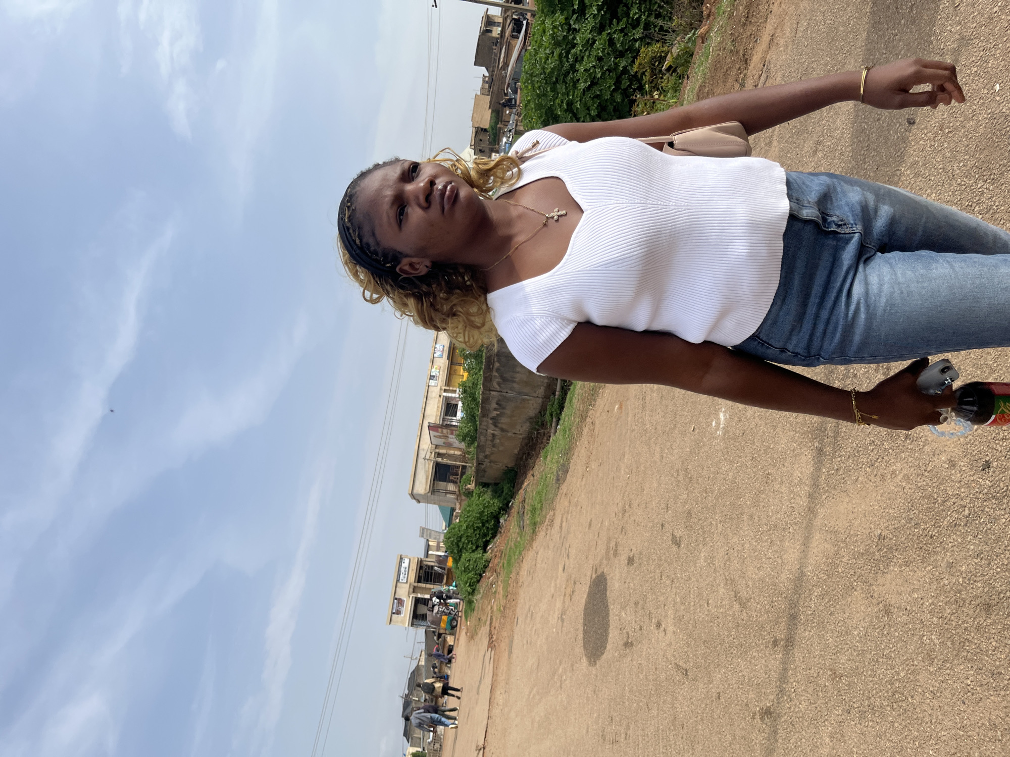 A woman with blonde-streaked hair, wearing a white ribbed top and blue jeans, walks on a dirt road in Rafin Par, Nigeria. She carries a dark bottle and a cell phone in her right hand, and a light-colored purse is slung over her left shoulder. In the background, several small buildings with shops line the road, and a three-wheeled vehicle is parked. Other people are visible walking in the distance. The sky is partly cloudy.