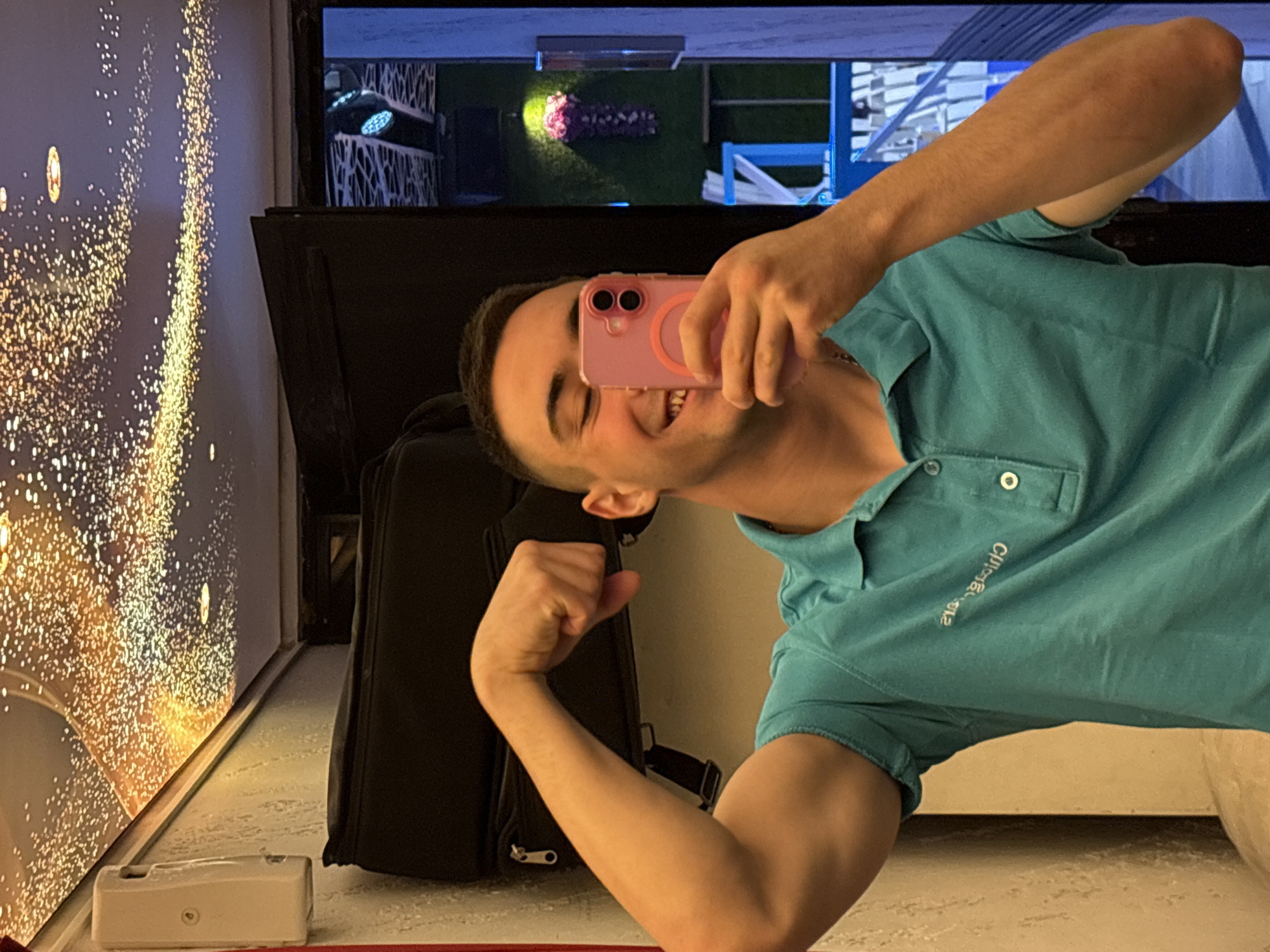 A man is taking a selfie indoors, likely in a hotel in Plovdiv, Bulgaria. He is wearing a turquoise polo shirt and holding a pink phone. He is smiling and flexing his arm. Behind him is a large black suitcase, a wall with a decorative pattern of gold dots, and a television screen showing a view of what appears to be a garden or outdoor area. The orientation of the image is rotated sideways.