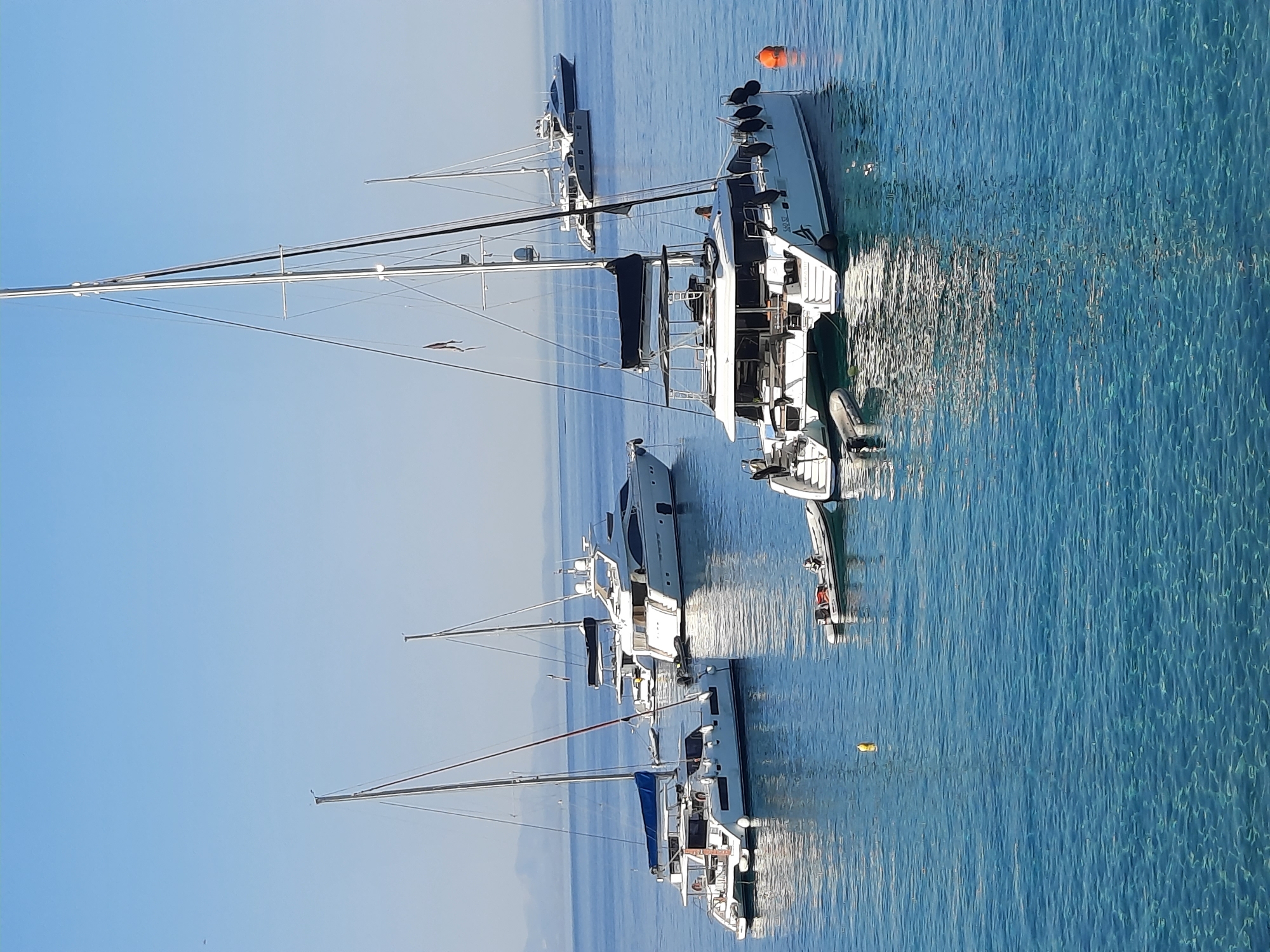 The image shows a fleet of yachts anchored in the clear blue waters of Platys Gialos, Greece. In the foreground, a white catamaran with two hulls is prominently featured. It has a large, white superstructure, black bimini top, and is equipped with a dinghy attached to its stern. A person is visible on the deck of this catamaran. To the left of the catamaran, another white catamaran is anchored, with a blue sail furled on its mast. Further in the background, two larger white motor yachts are visible, one with a sleek, modern design and the other more classic in appearance. In the distance, the hazy outline of mountains can be seen against the clear blue sky. The water is exceptionally clear, allowing the seabed to be faintly visible in some areas. The scene is peaceful, with the yachts at rest in a calm bay.