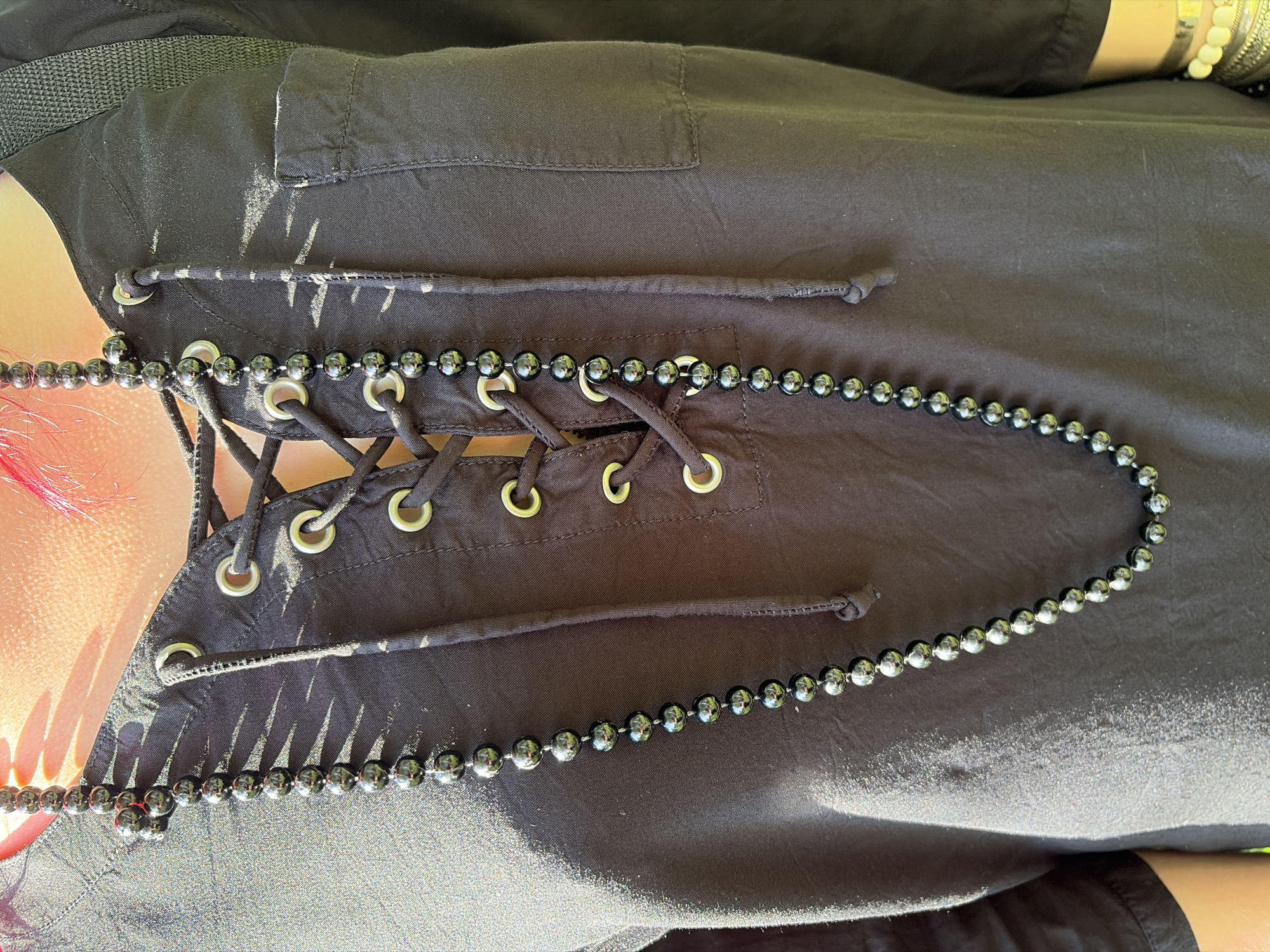 A close-up shot shows the chest area of a person wearing a black shirt with a lace-up front and a long black beaded necklace. The shirt has silver eyelets with black lacing. The background is blurred, suggesting an outdoor environment. There is no identifiable information about the location in Chelyabinsk, Russia, or specific actions.