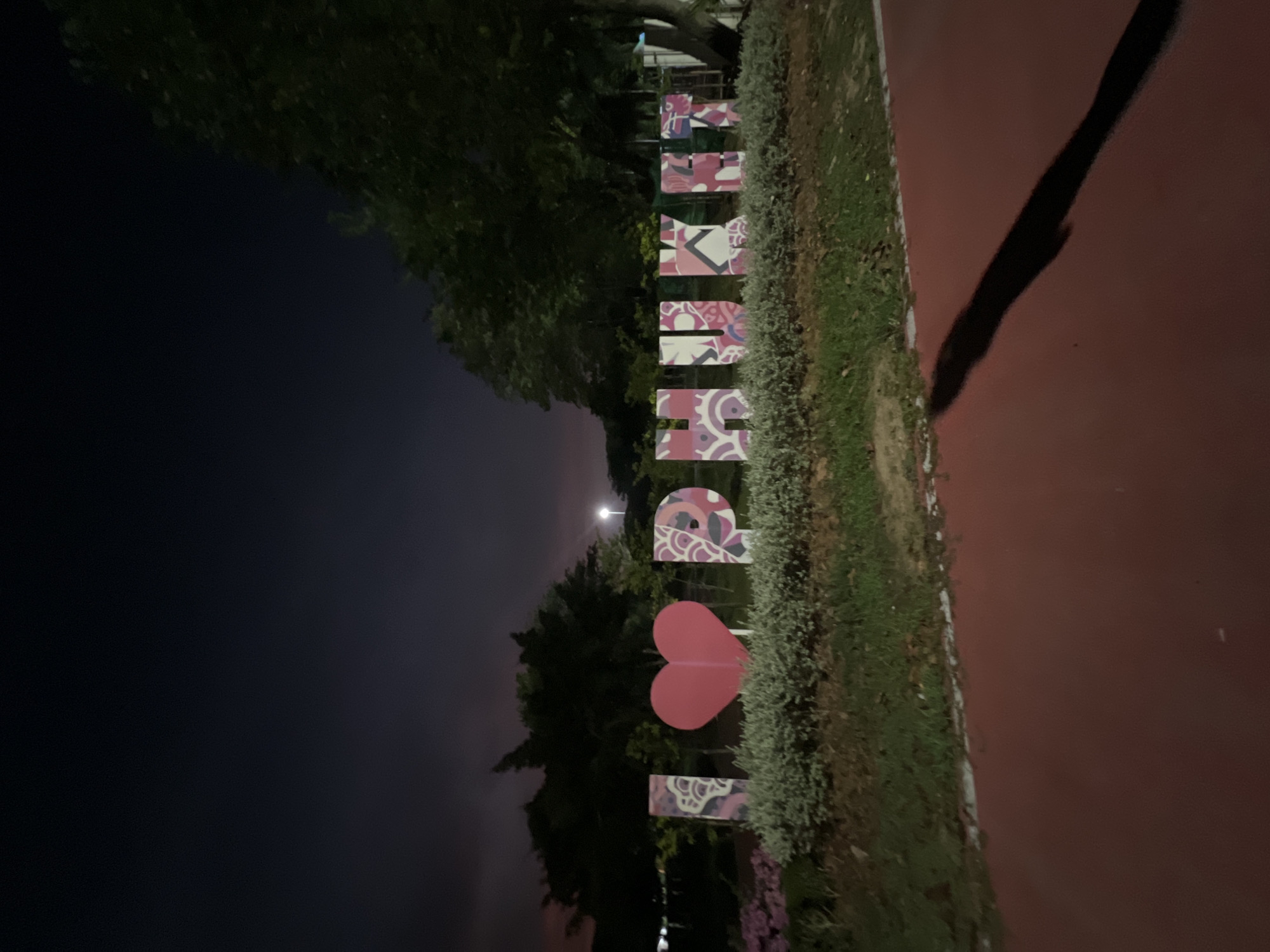 The image depicts an "I LOVE PHUKET" sign at night, located in Phuket City Municipality, Thailand. The letters of the sign are decorated with pink, purple, and white patterns. A red heart represents the word "LOVE". To the left of the sign are trees and a dark night sky. To the right, there is a strip of grass and a red-paved path. A long shadow is visible on the path.