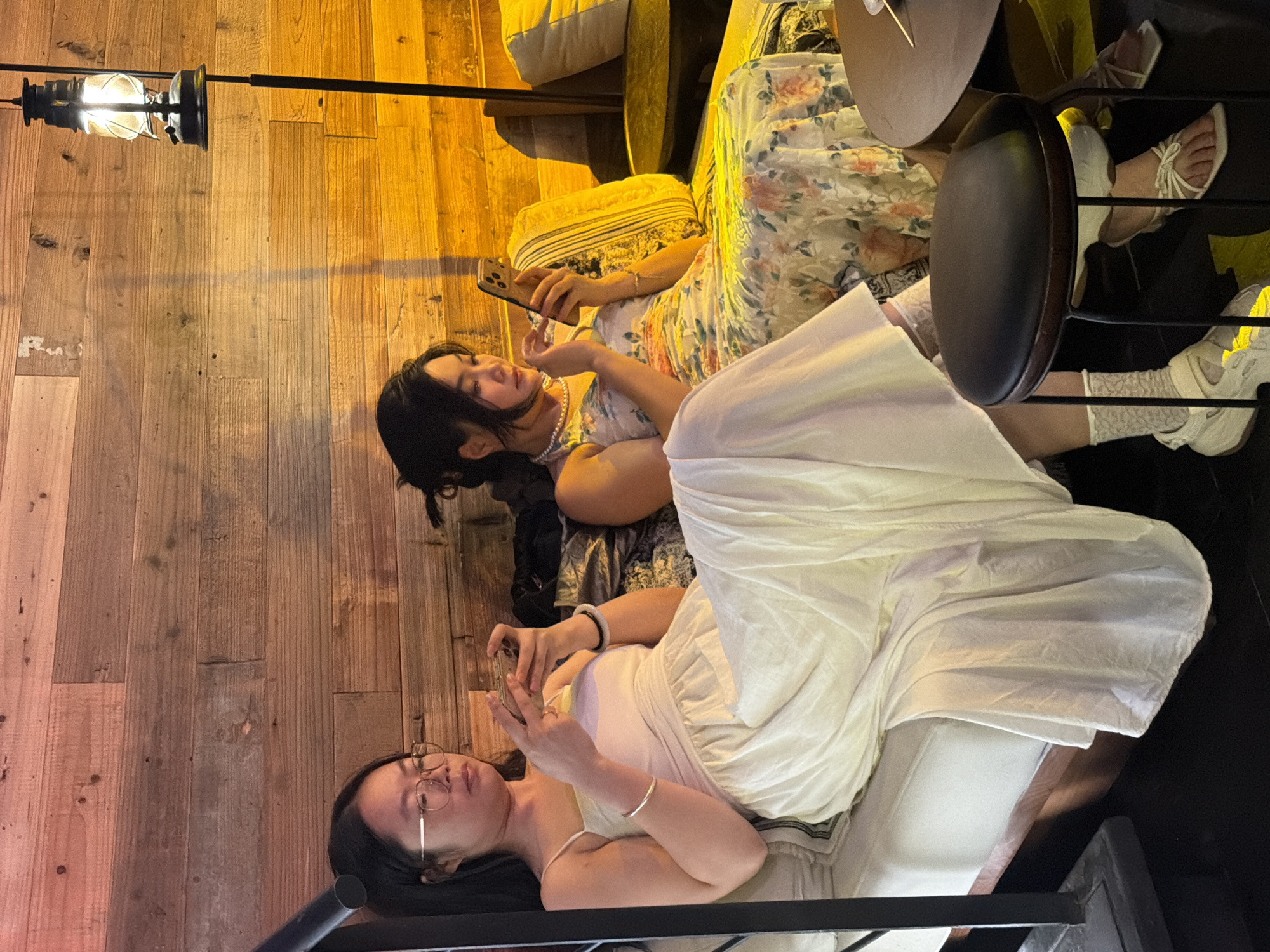 Two young women are depicted indoors, engrossed in their smartphones, likely in a cafe or lounge setting in 江北城街道, China. The image is rotated 90 degrees counter-clockwise, showing the subjects lying down or reclined against wooden paneling.
The woman at the top of the frame has dark hair and is wearing a floral patterned dress and a pearl necklace. She holds a smartphone in her right hand, gazing at its screen. Her right foot, clad in a white strappy sandal, is visible in the upper right quadrant of the image, suggesting she is seated with her feet up.
The woman at the bottom of the frame has dark hair, glasses, and is wearing a light-colored (possibly white or cream) dress or top and skirt combination. She also holds a smartphone, focusing on its display. A silver bracelet is on her left wrist, and a black and white bracelet is on her right. Partially visible on her feet are white sneakers worn with white lace-patterned socks.
The background features warm, rustic wooden paneling that covers the walls. An antique-style lantern with a glowing bulb hangs from a black metal bar on the wall in the upper left. Cushions or pillows are visible behind the women, suggesting comfortable seating. A dark, circular object (possibly a cymbal or a stool back) is positioned between the two women. The overall lighting is warm and dim, indicating an evening or nighttime setting. No visible text is present in the image.