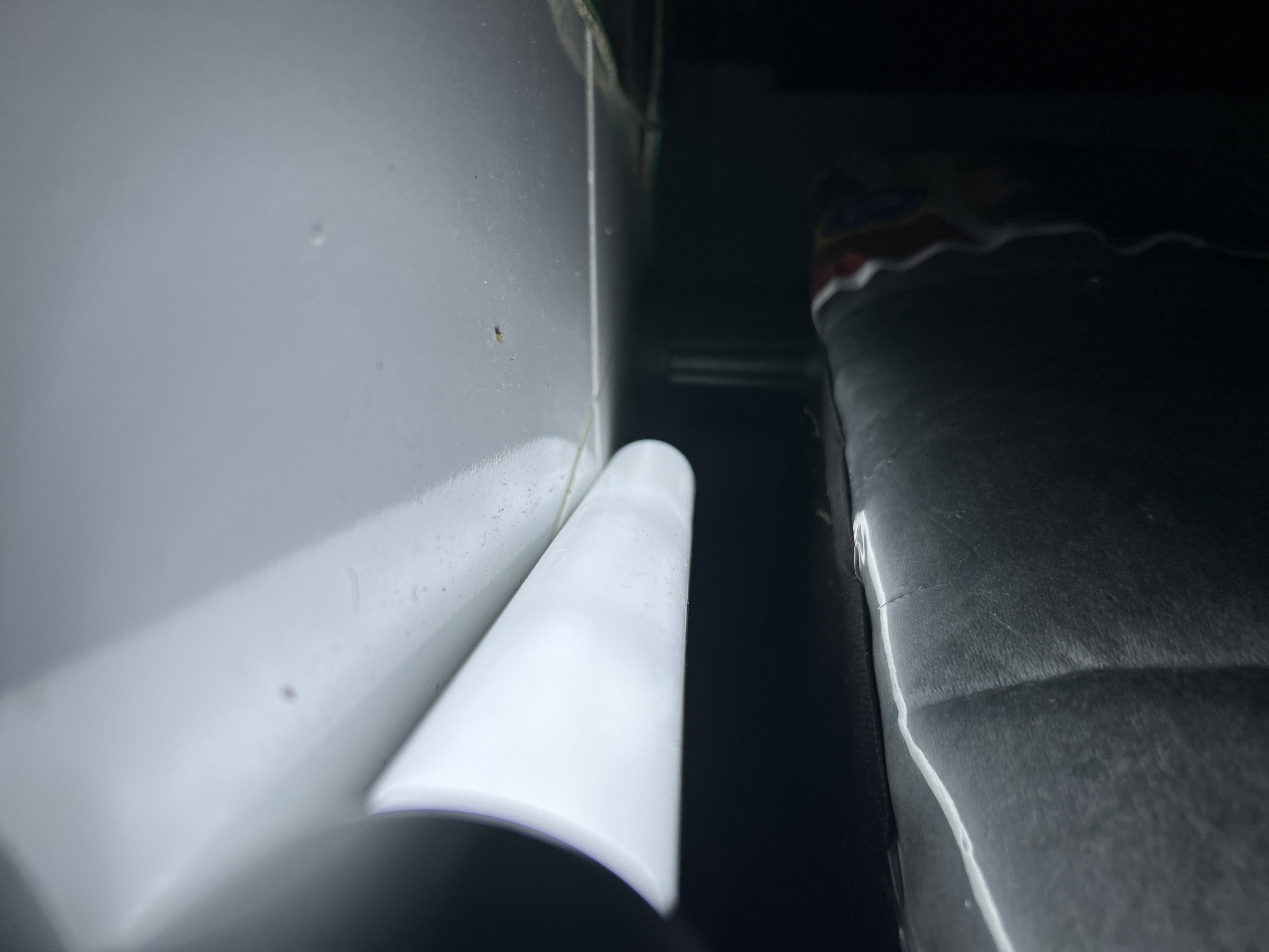 The image presents a close-up view of an interior space, characterized by a tight crevice between a white, possibly metallic, surface and a dark, textured surface. A cylindrical white object is partially visible in the foreground, its origin unclear due to the limited field of view. The overall lighting is dim, casting deep shadows and obscuring details in the background. The presence of the red, white, and blue pattern suggests the presence of bedding in the background, and the dark texture implies a seat or cushion. The image presents no evidence of the location being the city of Lhokseumawe, Indonesia.