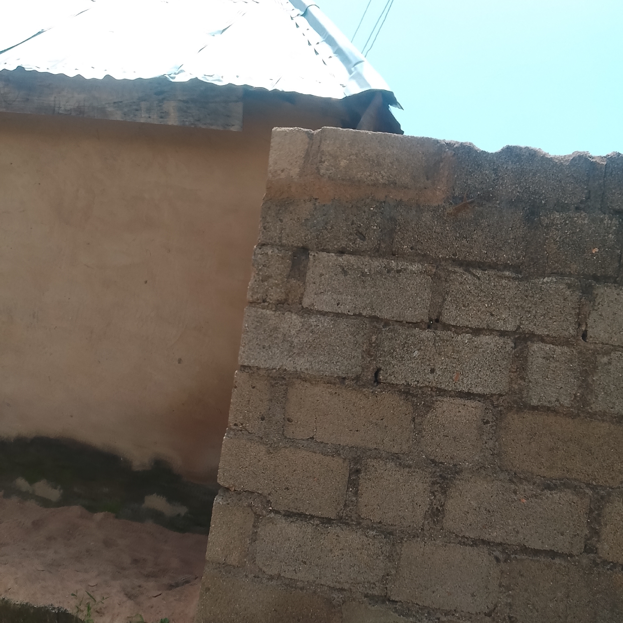 This media file depicts an outdoor scene in Jalingo, Nigeria, focusing on the exterior of a building. The dominant subjects are architectural elements: a rough, unfinished brick wall in the foreground and a section of a building with a corrugated metal roof in the background. The brick wall is constructed from rough, grey concrete blocks, showing the texture of the aggregate. The building in the background has a tan, plastered wall, with a dark, damp-looking band running horizontally across its lower visible section. The corrugated metal roof appears weathered and silvery, with visible seams and ridges.
The scene is set during the daytime, indicated by the bright, clear blue sky. The lighting suggests it could be mid-morning or early afternoon. There are no people visible in the frame, and no specific activity or event is taking place; it is a static architectural study.
Notable details include the unfinished nature of the brick wall, the likely presence of moisture or staining on the lower part of the building's wall, and the general impression of a practical, perhaps developing, construction site. The sky is a bright, unclouded blue, typical of sunny weather. The location cues, like the building style and the material choices, are consistent with a tropical or sub-tropical environment. There is no visible text within the image.