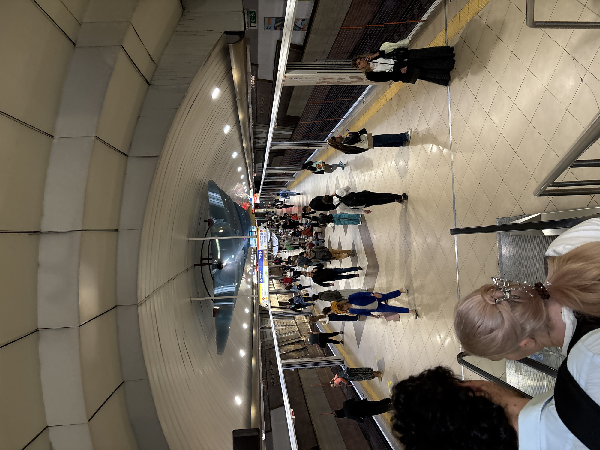 This image depicts a bustling underground metro station in Sofia, Bulgaria, captured from an elevated viewpoint, likely from an escalator. The station features a distinctive arched tunnel ceiling constructed from segmented, light-colored panels, illuminated by rows of recessed lights.
Numerous people are visible throughout the scene, engaged in commuting activities. Many individuals stand on the tiled platform, waiting for a train, walking, or engrossed in their phones. In the immediate foreground, a person with light-colored hair, adorned with a decorative hair clip, wearing a white shirt and dark vest, is seen ascending an escalator. Another person with dark, curly hair is just ahead of them.
The platform edge is marked by a yellow tactile paving strip. Various informational signs and advertisements line the platform. A large, colorful vertical sign provides metro line information and symbols, with visible Bulgarian text including "МЕТРОПОЛИТЕН" (Metropolitan) and "БАНКИ" (Banks). A banner prominently displays the city name "SOFIA," confirming the location. Other visible text includes an "exit" symbol and "ОБУВКИ" (Shoes), likely an advertisement.
The overall impression is that of a modern, well-lit, and functional urban transit hub during what appears to be a busy period, with commuters awaiting their next train.