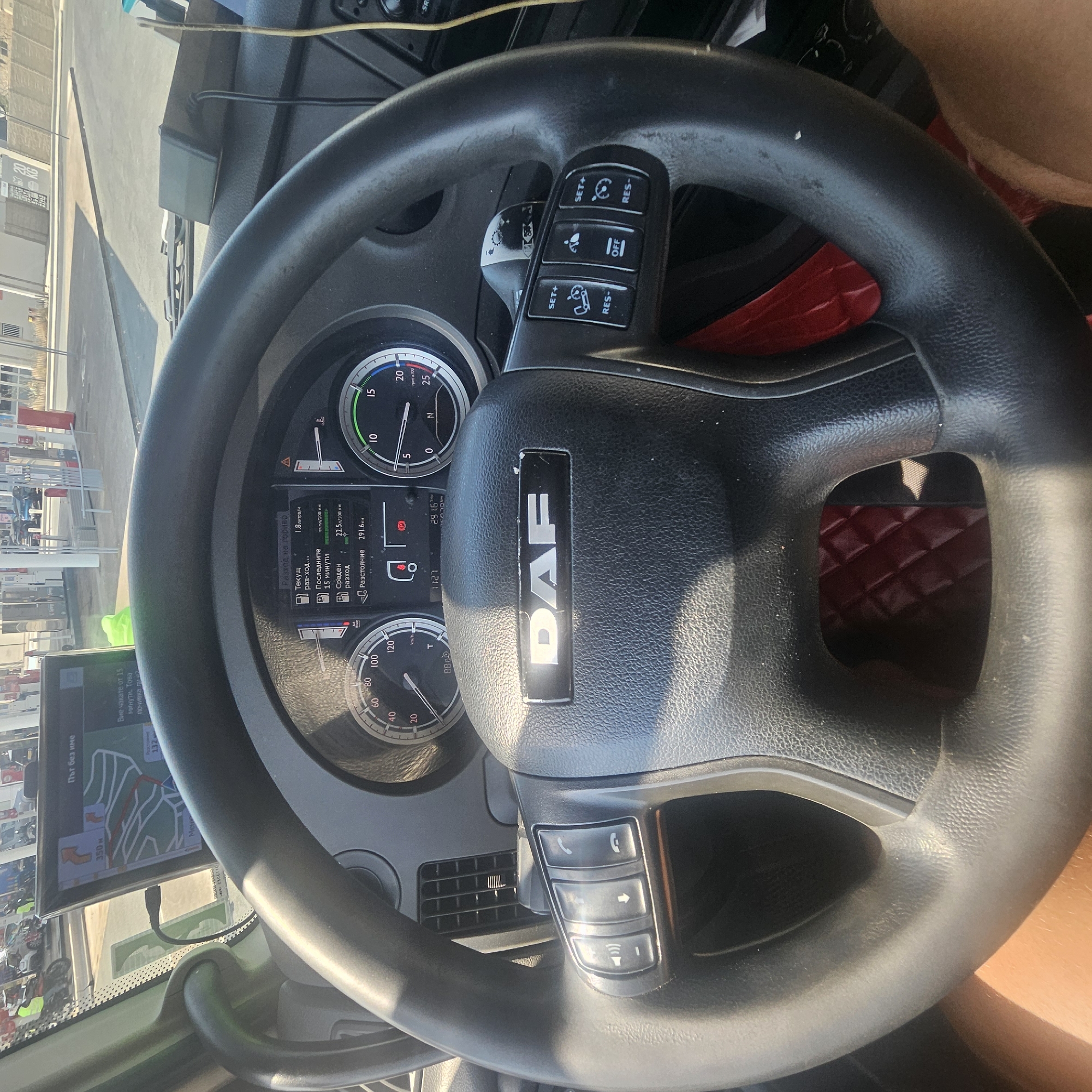 The image shows the interior of a DAF truck cab, viewed from the driver's perspective. The steering wheel, bearing the DAF logo, is prominent in the foreground. The dashboard displays various gauges, including a speedometer and tachometer, and a digital screen showing fuel consumption data. On the left side of the steering wheel are media control buttons. On the right side are cruise control buttons. A GPS navigation device is mounted on the windshield. Outside the truck, a gas station with multiple pumps and vehicles is visible. The scene appears to be at a gas station in La Palme, France, as indicated by the "La Palme" text on the GPS screen. The lighting suggests daytime. No individuals are clearly visible within the cab.