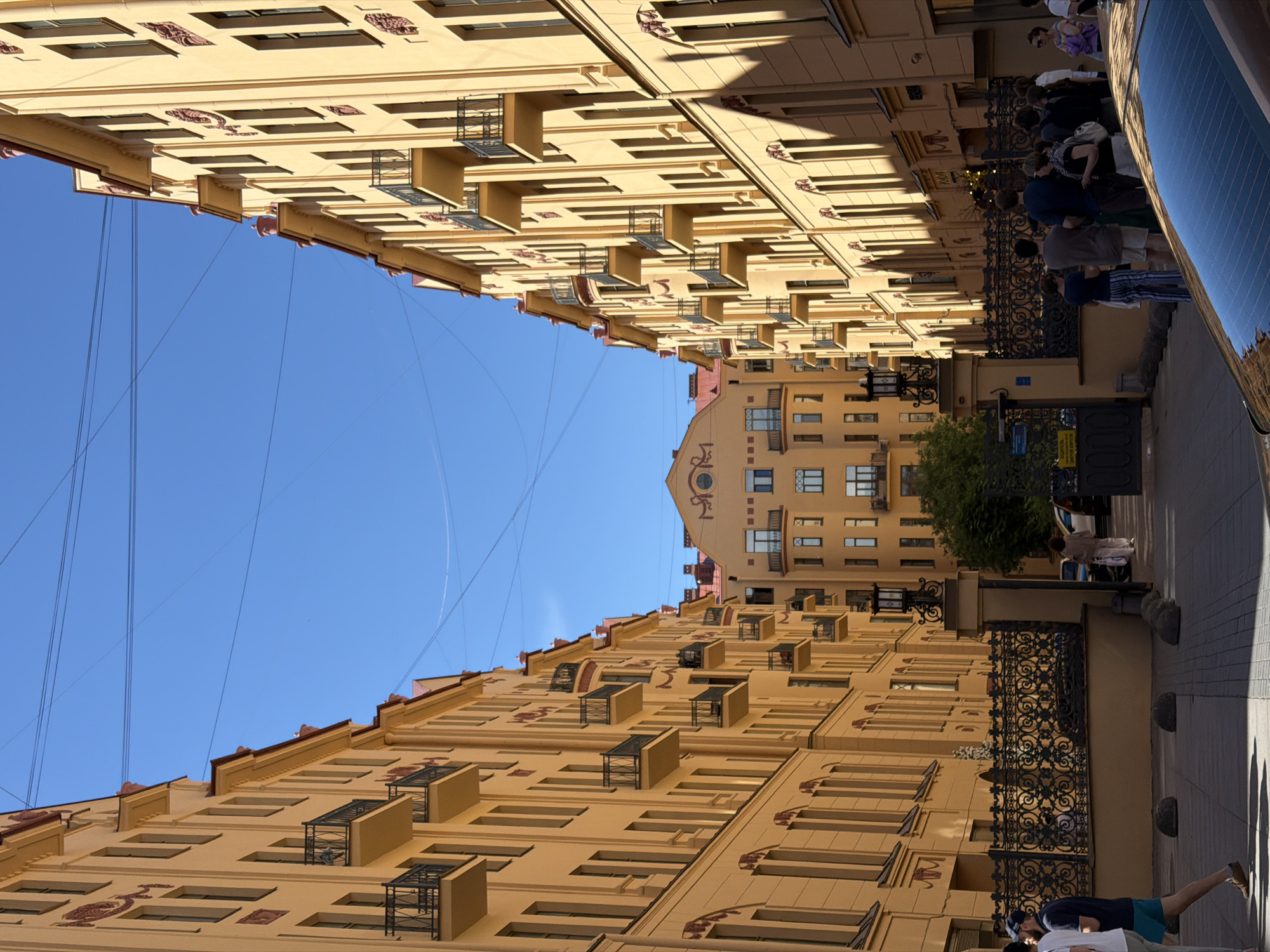 The image depicts a street or courtyard in Saint Petersburg, Russia, flanked by two multi-story buildings of pale yellow (ochre) with red-orange rooflines and decorative elements. These buildings, appearing symmetrical, feature numerous vertically aligned windows and multiple balconies with ornate black wrought-iron railings. A third, similarly colored building with a distinct peaked roof and a round window is visible at the end of the passage, framed by the two primary structures.
The sky is clear blue with several thin, dark electrical wires crisscrossing overhead. Bright sunlight casts distinct shadows on the ground and parts of the buildings.
At street level in the foreground, a decorative black wrought-iron fence on a low stone wall separates the immediate foreground from an inner courtyard or further passage. An ornate black lamp post stands on a stone pillar to the right of the central passage. Several round stone bollards are arranged on the paved walkway in the foreground.
Multiple individuals are present on both sides of the passage. On the left, a man in a dark blue t-shirt and light green shorts walks towards the viewer. On the right, a group of people is gathered near the fence, some facing into the passage, others conversing. Their attire is casual summer wear. Further back in the passage, near a patch of green foliage, a woman in light-colored clothing walks away from the viewer. A car's rear window, reflecting the sky and power lines, is partially visible in the bottom right corner of the frame.