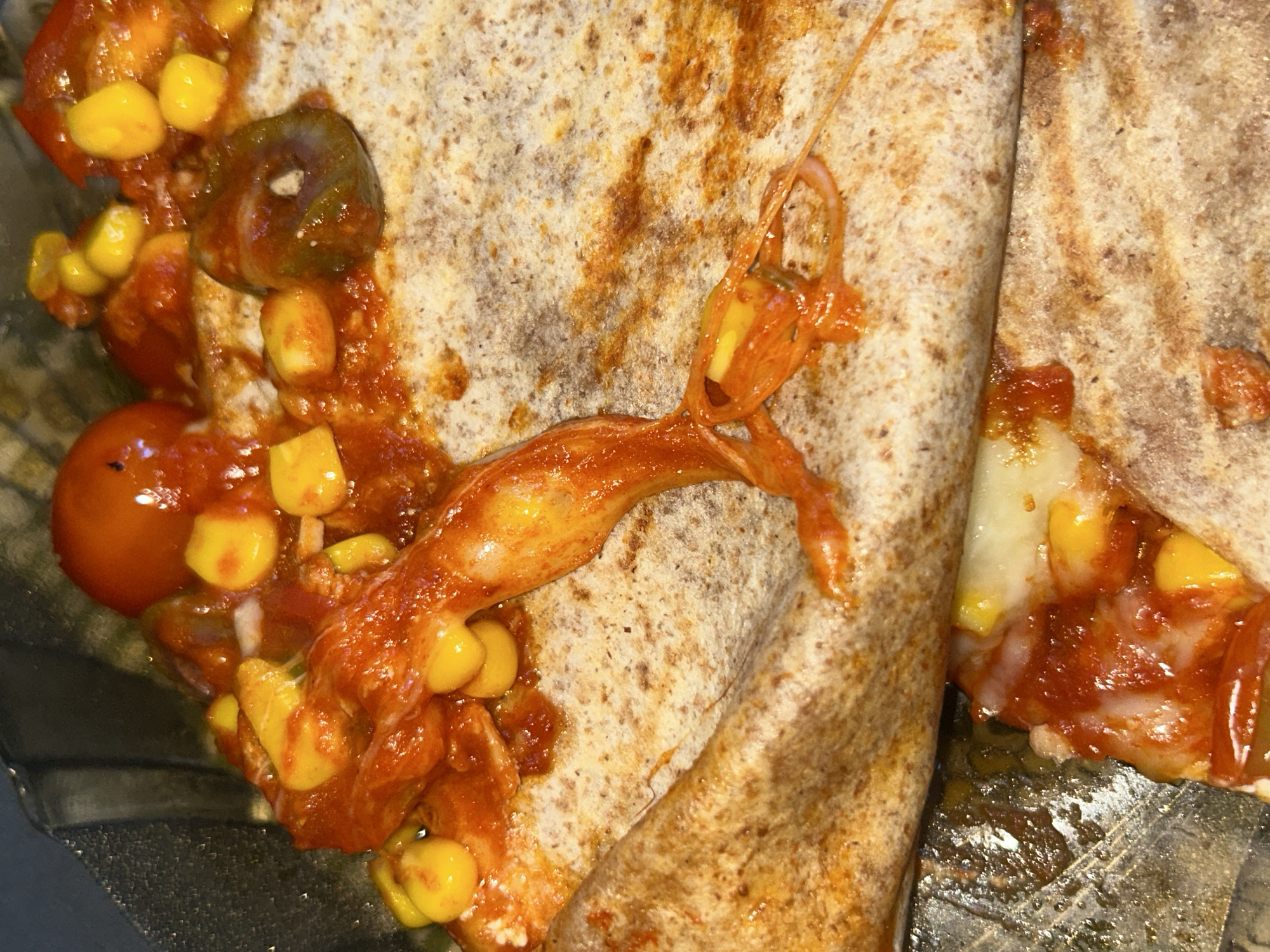 The image shows a close-up, overhead view of a quesadilla, cut in half, inside a clear plastic container. The quesadilla is filled with melted cheese, corn kernels, chopped tomatoes, and pieces of what appear to be jalapeno peppers. The tortilla is golden brown and slightly charred in places. The filling is spilling out of the quesadilla in some areas, with cheese strings extending outward. The container is sitting on a dark, reflective surface. There is no information available to determine the location in the city of Sofia, Bulgaria.