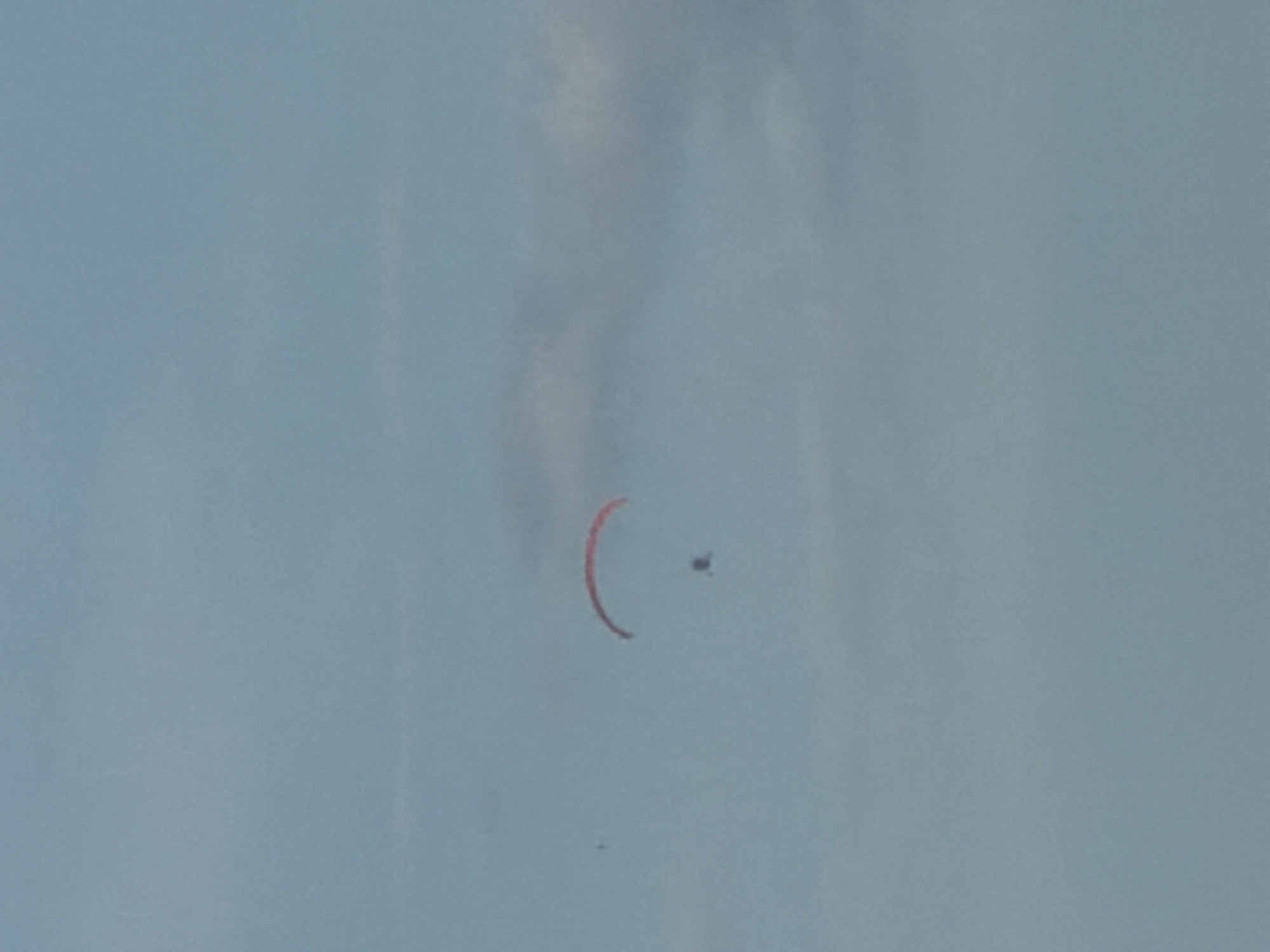 A paraglider is captured mid-flight against a hazy blue sky. The paraglider's wing is a vibrant red, forming a distinct arc. Below the wing, a small, dark figure, presumably the pilot, is suspended. The paraglider appears to be at a moderate altitude. The background consists of a pale blue sky with faint, wispy clouds. The image lacks any identifiable city landmarks, suggesting it was taken from an elevated perspective, possibly a helicopter or another paraglider, and does not contain enough information to pinpoint a specific location within Nha Trang, Vietnam.