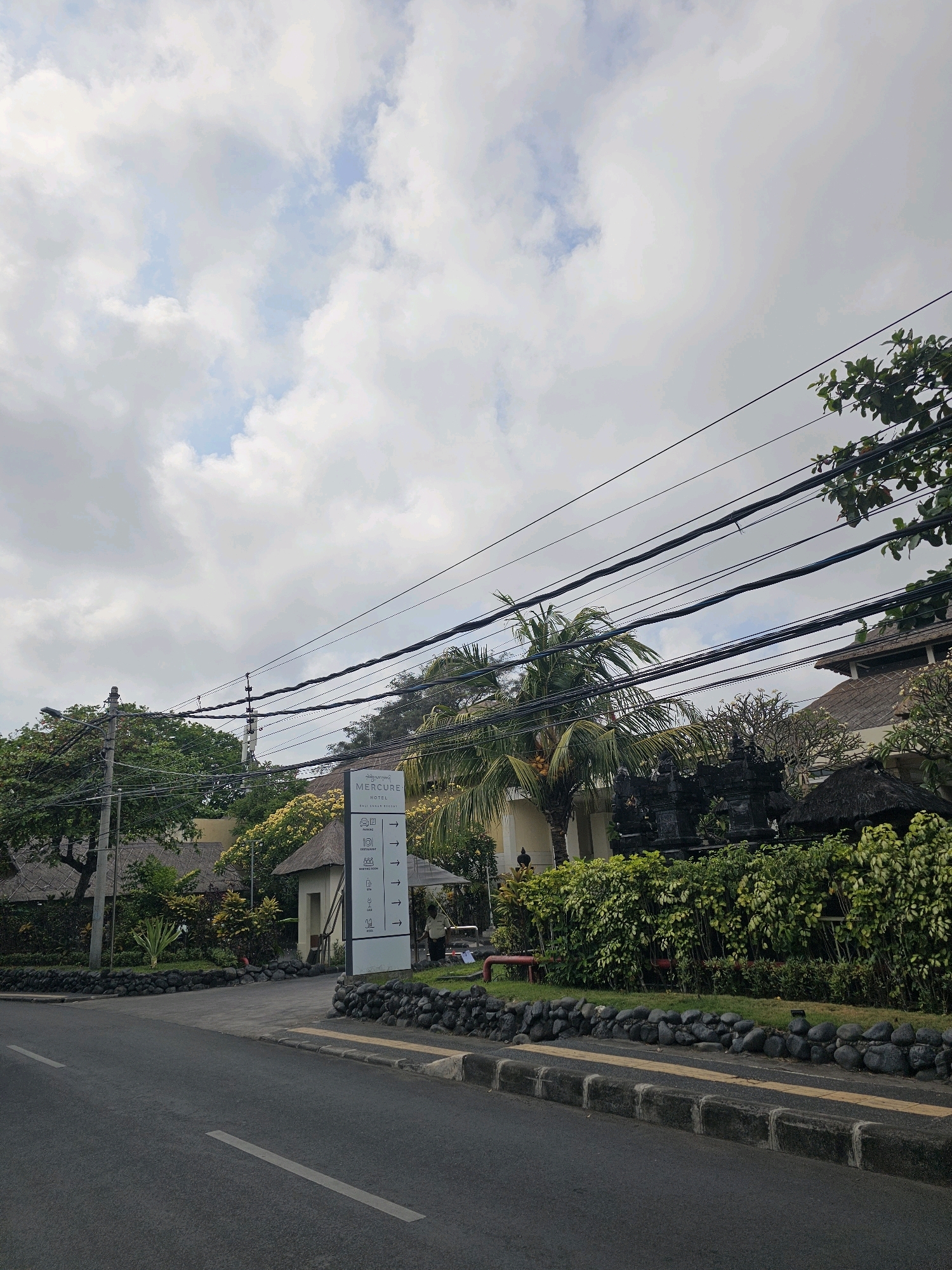 A wide, paved road curves to the left, with a gray asphalt surface and white lane markings. A concrete curb with a yellow tactile strip runs along the right side of the road. Beyond the curb is a low wall constructed of dark, rounded stones, topped with green grass. Behind the stone wall, lush green bushes and trees form a dense hedge.
To the right of the road, a tall white sign stands, bearing the logo and name "Mercure HOTEL" in black text. Below the hotel name are directional icons and text indicating various facilities such as "PARKING," "RESTAURANT," "GYM," and "MEETING ROOM," each with a directional arrow pointing to the right. A person in a light-colored uniform stands near the sign, facing away from the camera.
Behind the hedge and sign, a building with a thatched roof and white walls is visible, partially obscured by trees. A palm tree with yellow fruits stands to the left of the sign, its fronds reaching upwards. Several utility poles and a complex network of black power lines crisscross the sky above the scene. The sky is filled with a mix of white and gray clouds, with patches of blue visible. The overall impression is of a tropical, humid environment, likely in a tourist area. This scene is consistent with the entrance to the Mercure Resort Sanur in Denpasar, Indonesia, known for its tropical ambiance and proximity to the coast.