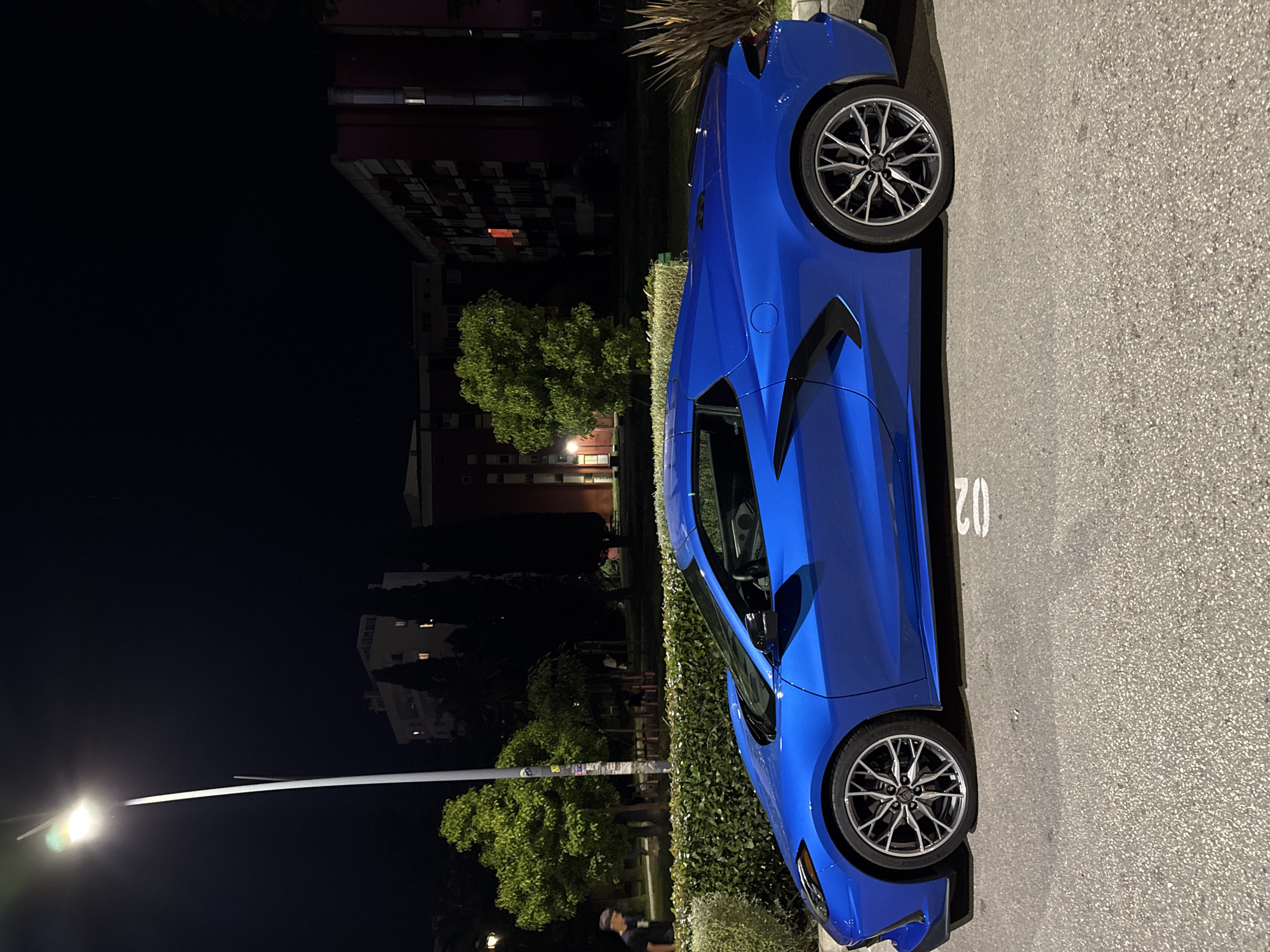 A blue sports car is parked on an asphalt surface in what appears to be a parking lot or roadside area. The number "02" is painted on the asphalt near the car. The scene is captured at night, with the surrounding environment lit by streetlights. There are trees and buildings visible in the background. Based on the architecture and the presence of Mediterranean vegetation, the location may be Budva, Montenegro. However, this would require additional supporting information. There is a person in the lower part of the image.