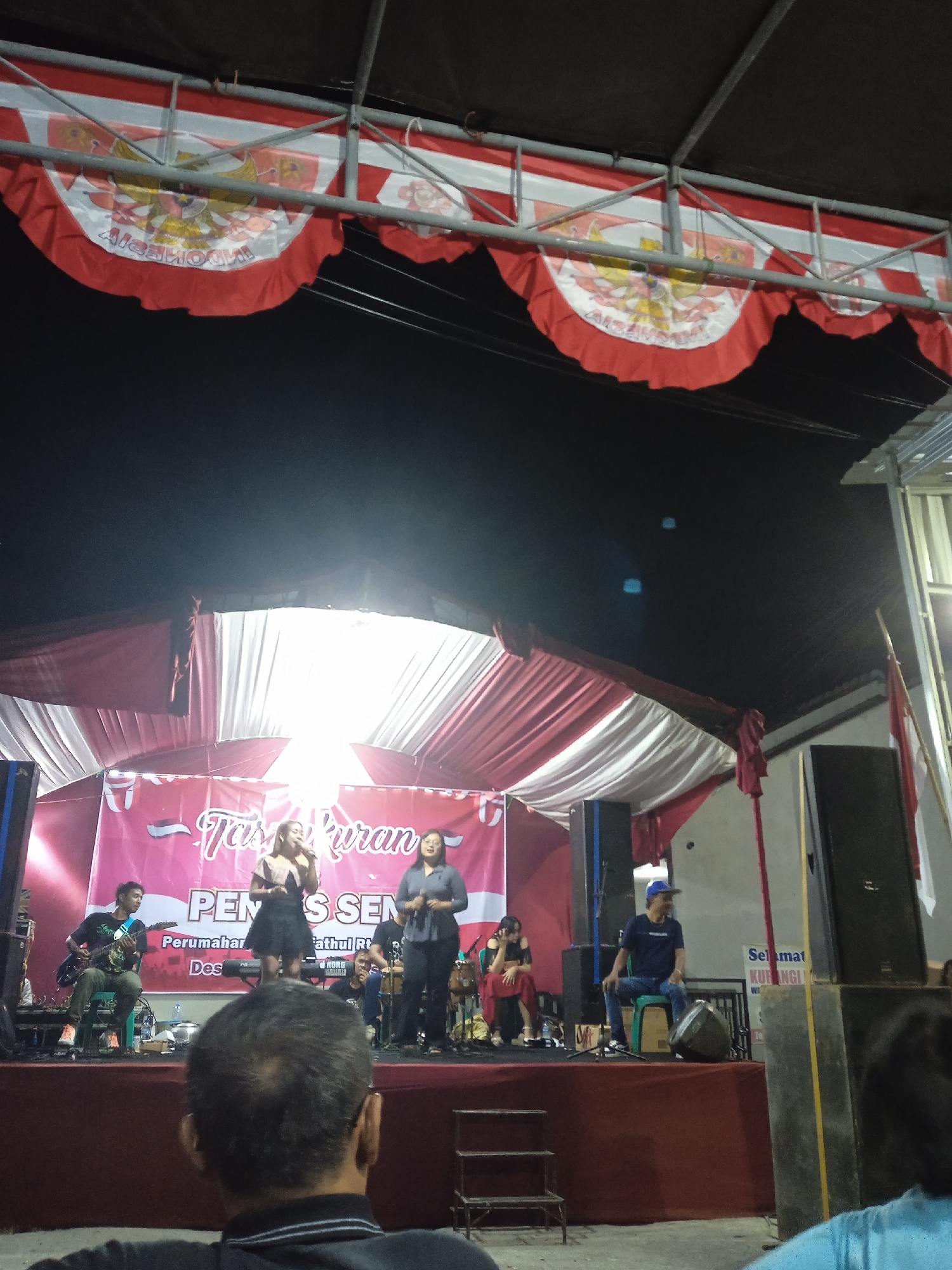 The image depicts an outdoor evening event, likely a performance or gathering, taking place in Cangkring, Indonesia. A stage is set up under a canopy decorated with red and white fabric and Indonesian flags, featuring the Garuda Pancasila emblem. On the stage, there are musical instruments, including a guitar and a keyboard, and several individuals. Two women are singing into microphones, while a man plays the guitar. Another man is seated on a chair on the right side of the stage. A banner behind the performers reads "TAS...KURAN PEN...S SEN...", with smaller text below indicating "Perumahan..." and "Des...". The audience is partially visible in the foreground, with one person's back to the camera. Large speakers are positioned on the sides of the stage. The overall atmosphere suggests a community event or celebration.