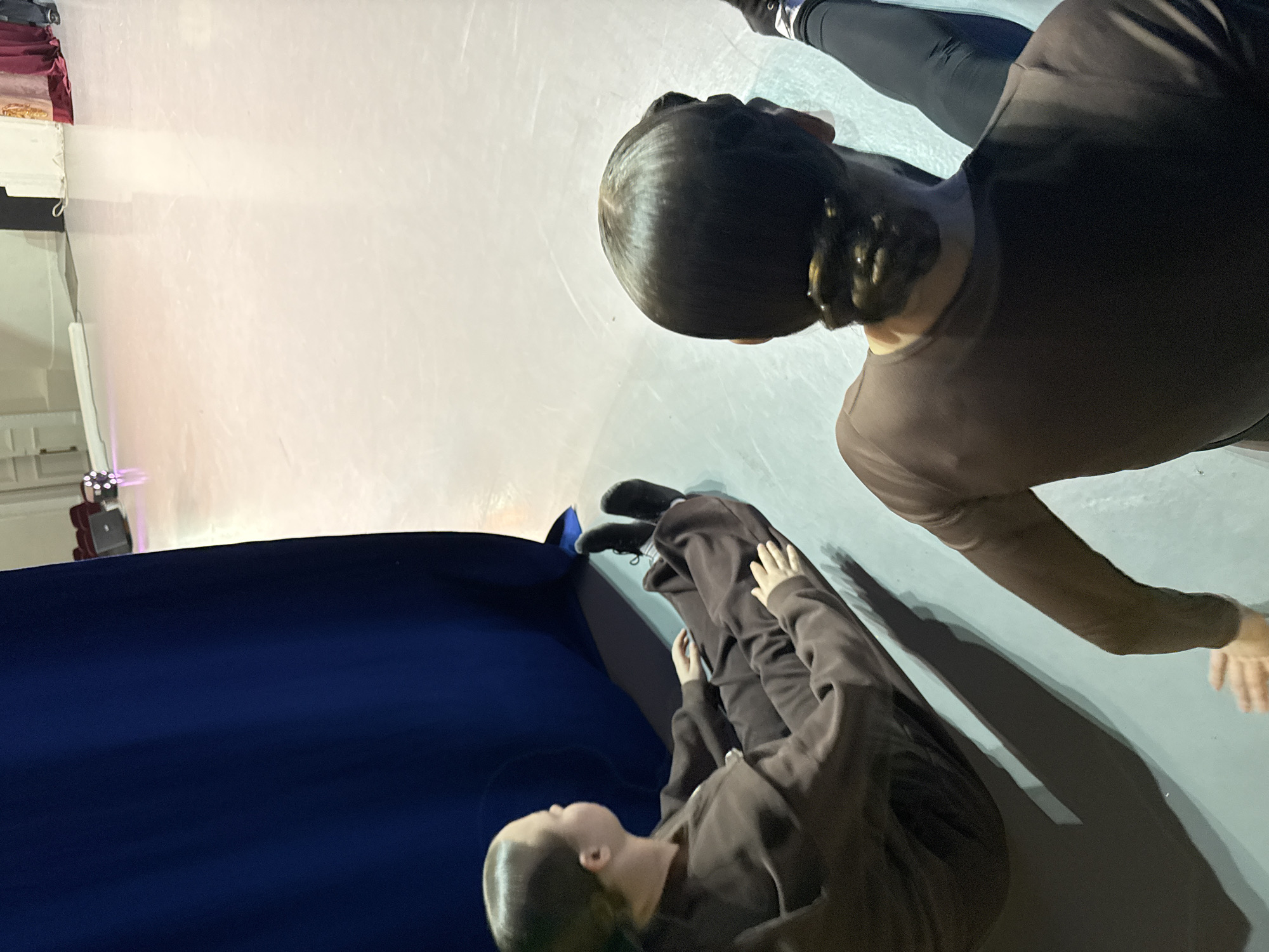 The image shows an indoor ice rink in Perm, Russia, where two figures are visible. One figure is standing on the ice, wearing dark leggings and a dark brown, long-sleeved shirt. The other figure is near a blue sheet, dressed in a dark brown outfit, and appears to be lying down on the ice or a surface near the rink. The ice rink is brightly lit with artificial lighting. A structure with a red decorative element is visible in the background.