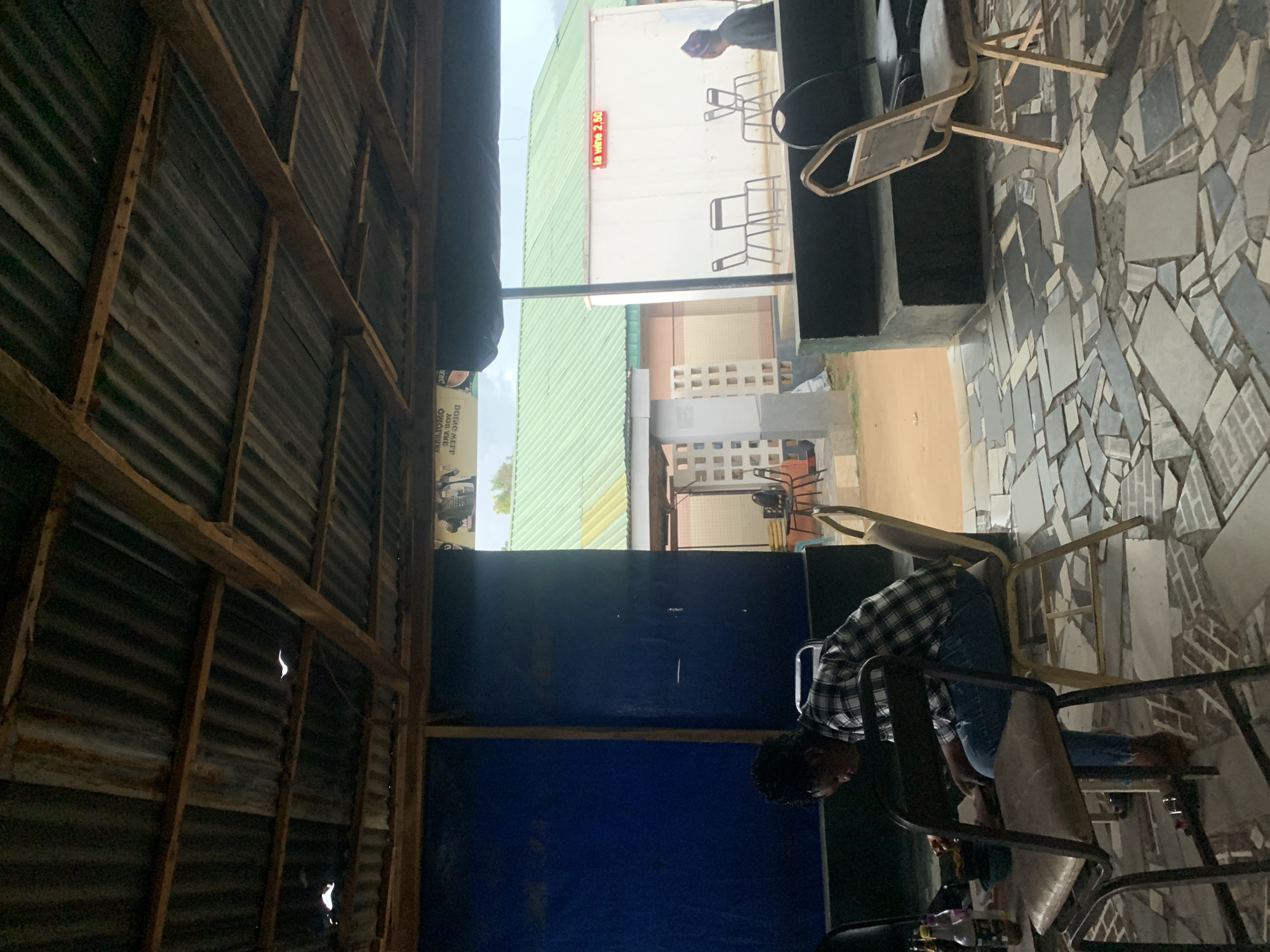 The image depicts an interior scene, likely within a restaurant or informal eating establishment in Kabong, Nigeria. A man in a plaid shirt and jeans is seated, possibly interacting with items on a table. The visible interior structure suggests a simple construction, with corrugated metal roofing and wooden supports. Outside, a building with "la wine 2.50" is visible. The floor is tiled with irregular pieces of light-colored tiles. Several chairs are scattered, seemingly positioned for patrons. Overall, the setting indicates a modest business operating in Kabong.