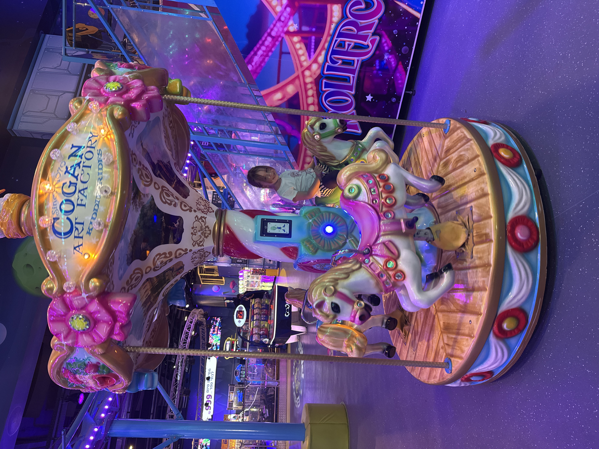 The image shows a carousel ride in a brightly lit indoor amusement park. A young child is riding one of the carousel's horses. The carousel has pink flower decorations at the top, and the side of the carousel says "Cogan Art Factory" and "Kiddie-Rides". A sign in the background reads "Ollifere", the location of the scene is Tel Aviv, Israel. Several arcade games and other amusement rides can be seen in the background. The floor is a speckled purple.