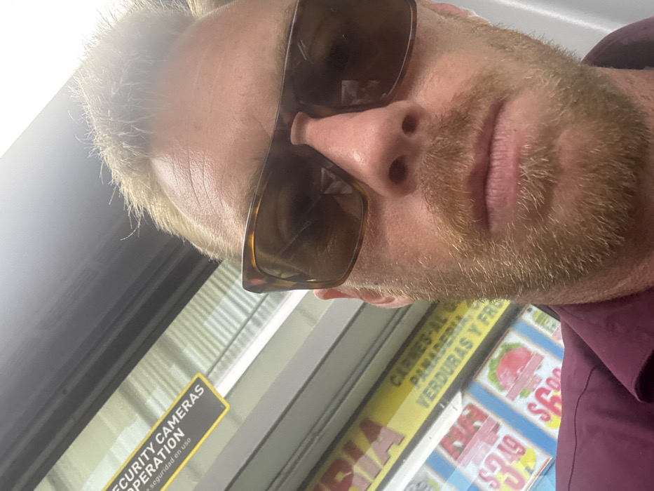 A man wearing sunglasses is close to the camera in a selfie. He has fair skin and a blonde beard. The background shows a sign reading "SECURITY CAMERAS OPERATION" with the words "seguridad en uso" underneath. Adjacent to this is a display advertising "CARNES-AL G" and "PANADERIA, VERDURAS Y FRO" with prices such as $3.49. The image is taken at an angle, with the man's face slightly tilted. He is wearing a purple shirt.